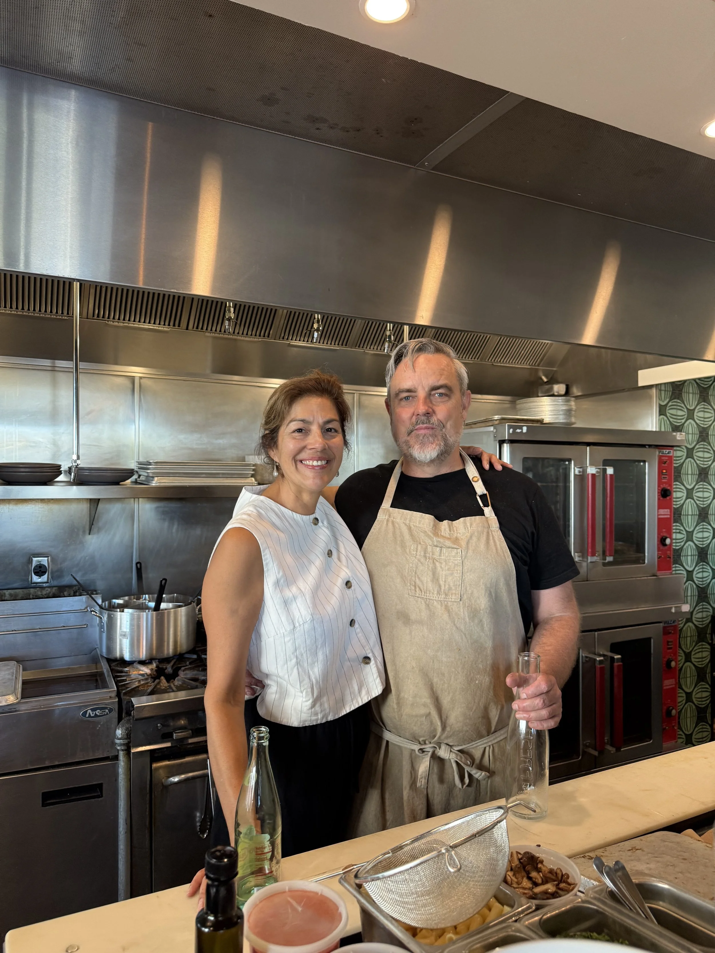 A smiling woman and a serious man wearing aprons standing in a professional kitchen with cooking equipment and food on the counter.