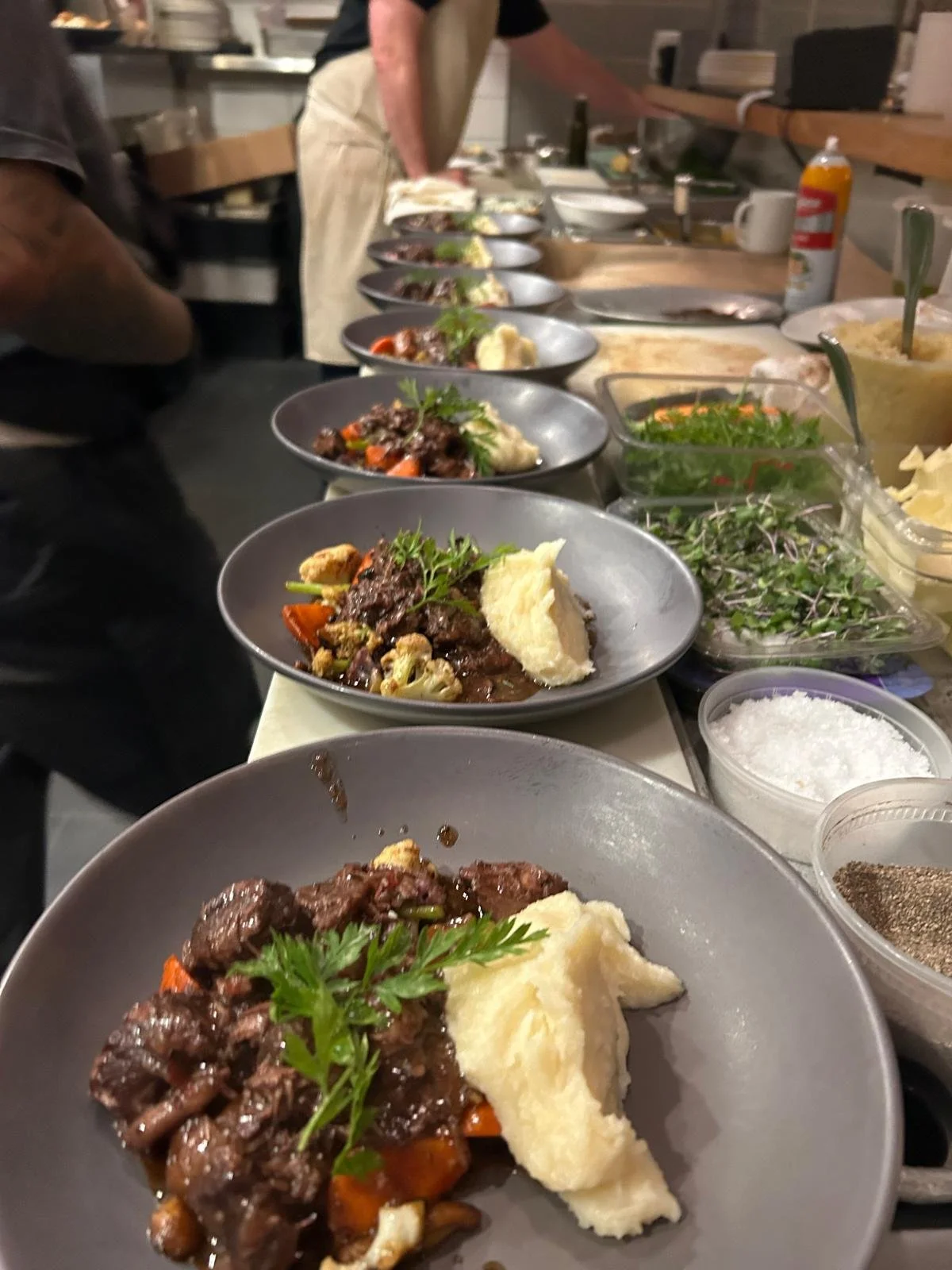A line of plated beef stew with mashed potatoes, garnished with parsley, on a kitchen counter. There are various ingredients, utensils, and garnishes nearby, with a person preparing food in the background.