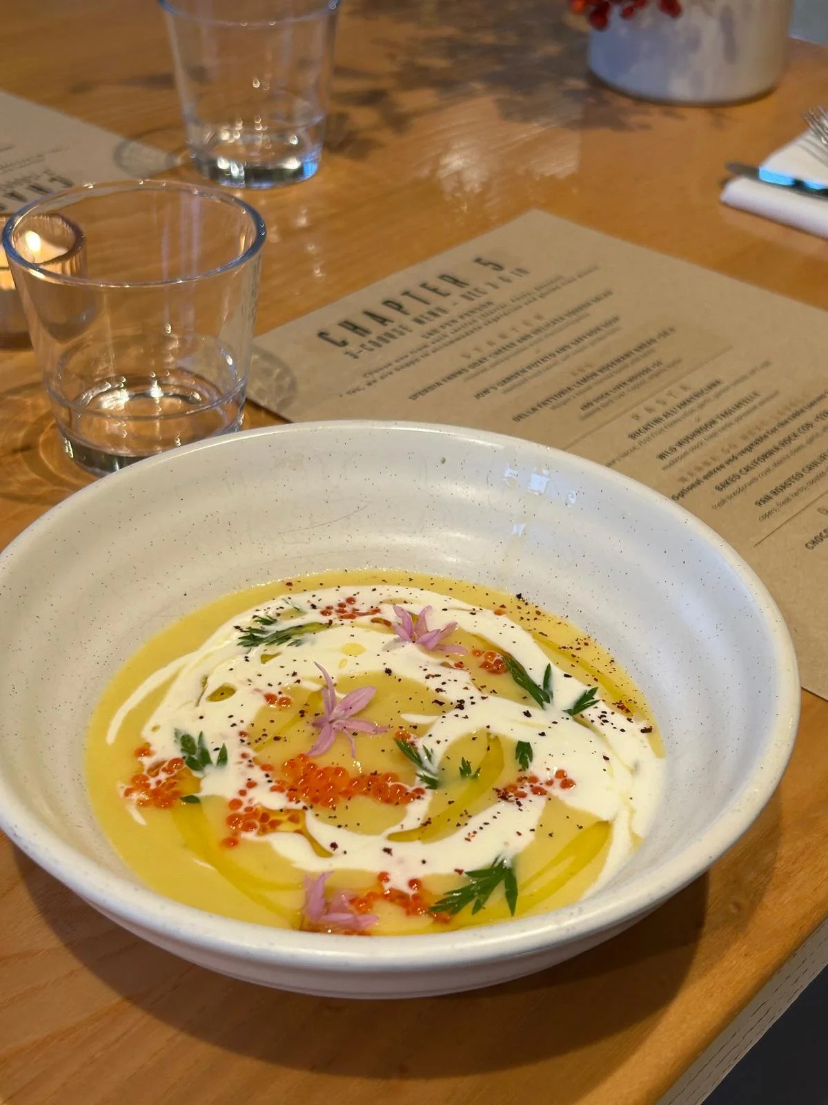 A bowl of creamy white soup garnished with pink edible flowers, green herbs, black pepper, and red-orange fish roe, served on a wooden table with a restaurant menu, glasses of water, and utensils in the background.