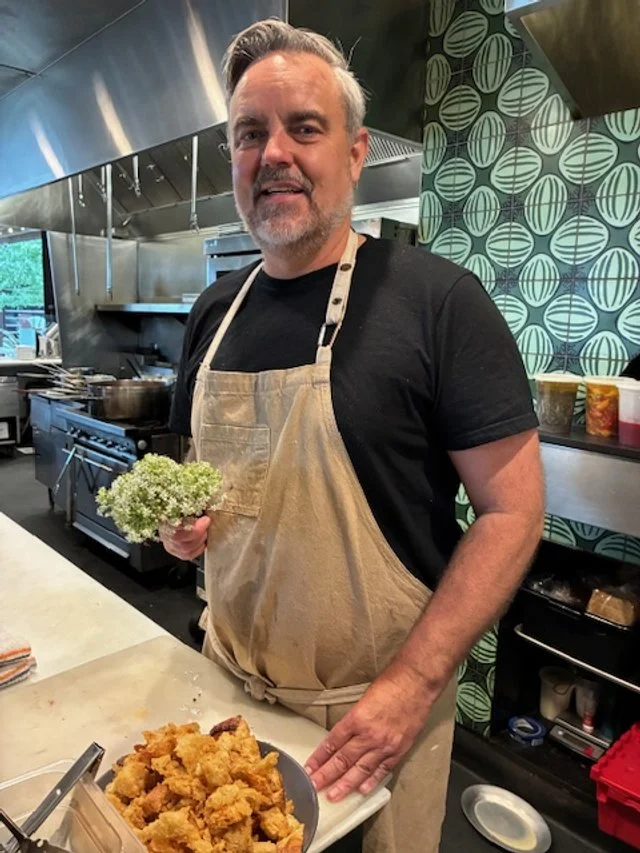 A man with gray hair and a beard wearing a black t-shirt and beige apron holding a bunch of green herbs in a restaurant kitchen with stainless steel appliances and patterned green wallpaper, standing near a tray of fried food.