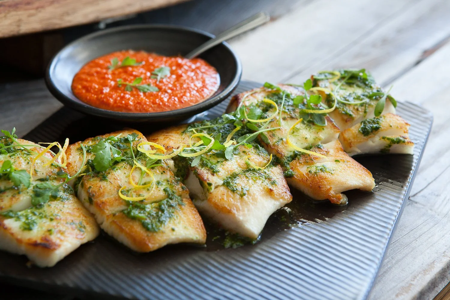 Grilled fish fillets garnished with greens and lemon zest, served with a side of red dipping sauce with herbs.