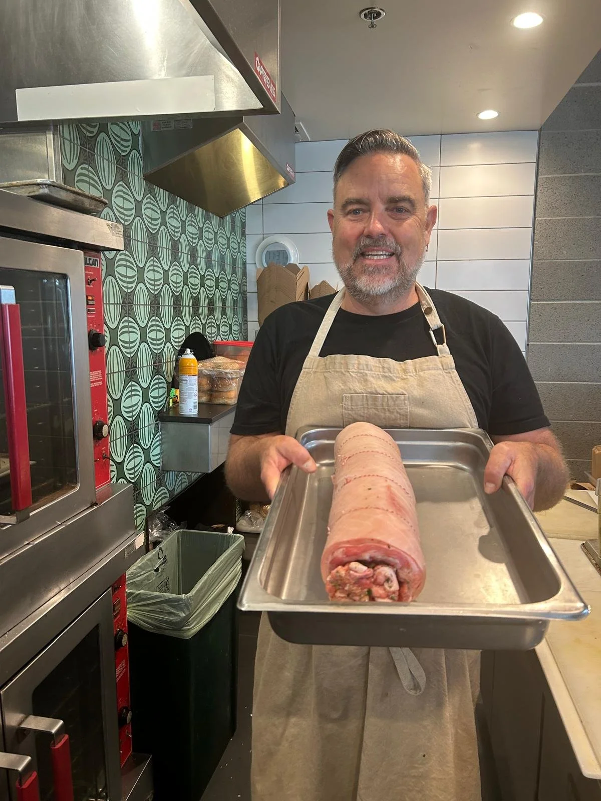 A man with a beard and short dark hair, wearing a beige apron, holding a tray with a rolled beef tenderloin roast, smiling at the camera in a kitchen.
