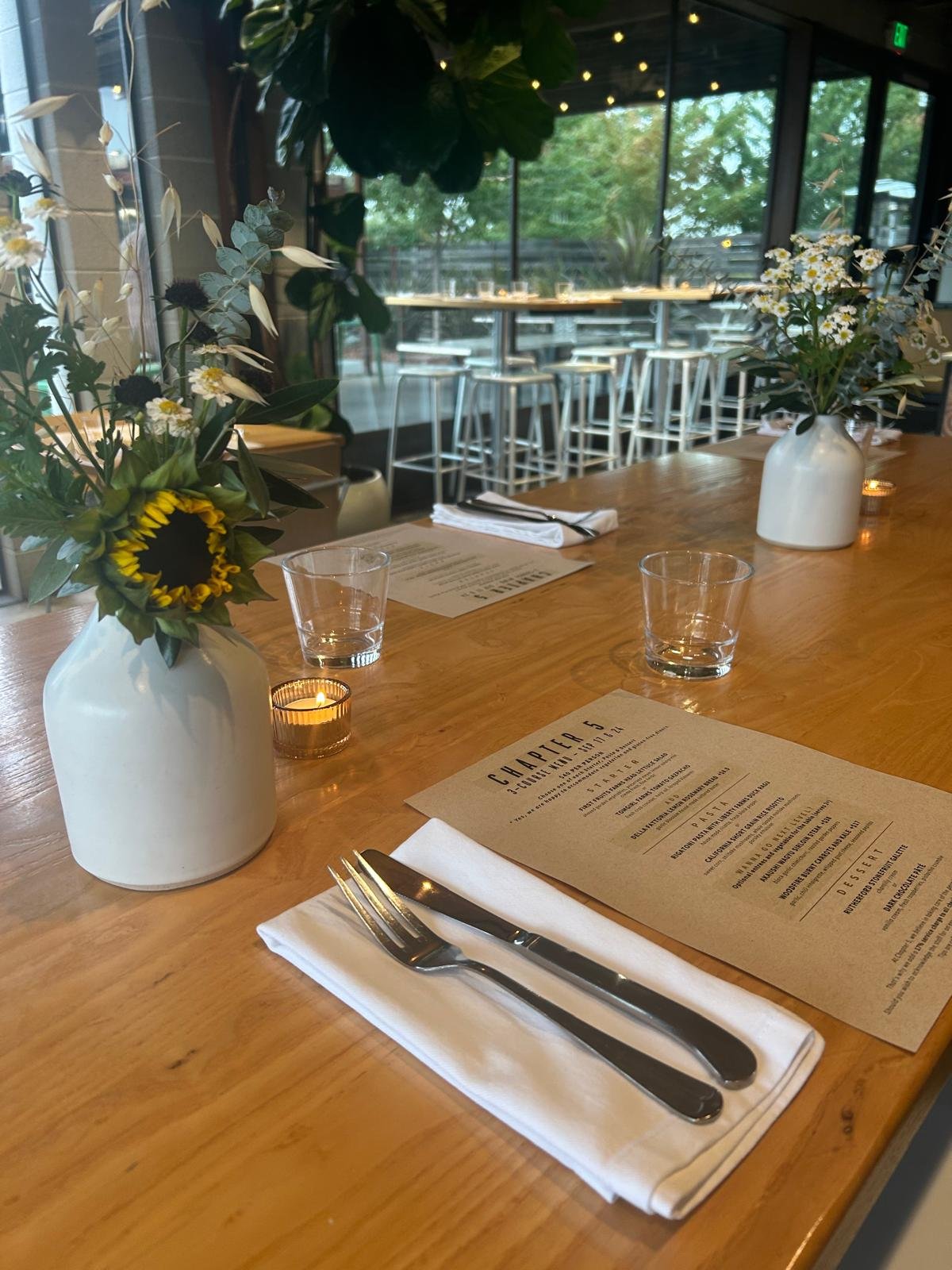 Dining table set with a white napkin, fork, and knife, two flower vases with white and yellow flowers, two small candles, and a menu in a modern restaurant with large glass windows and a view of outdoor seating.