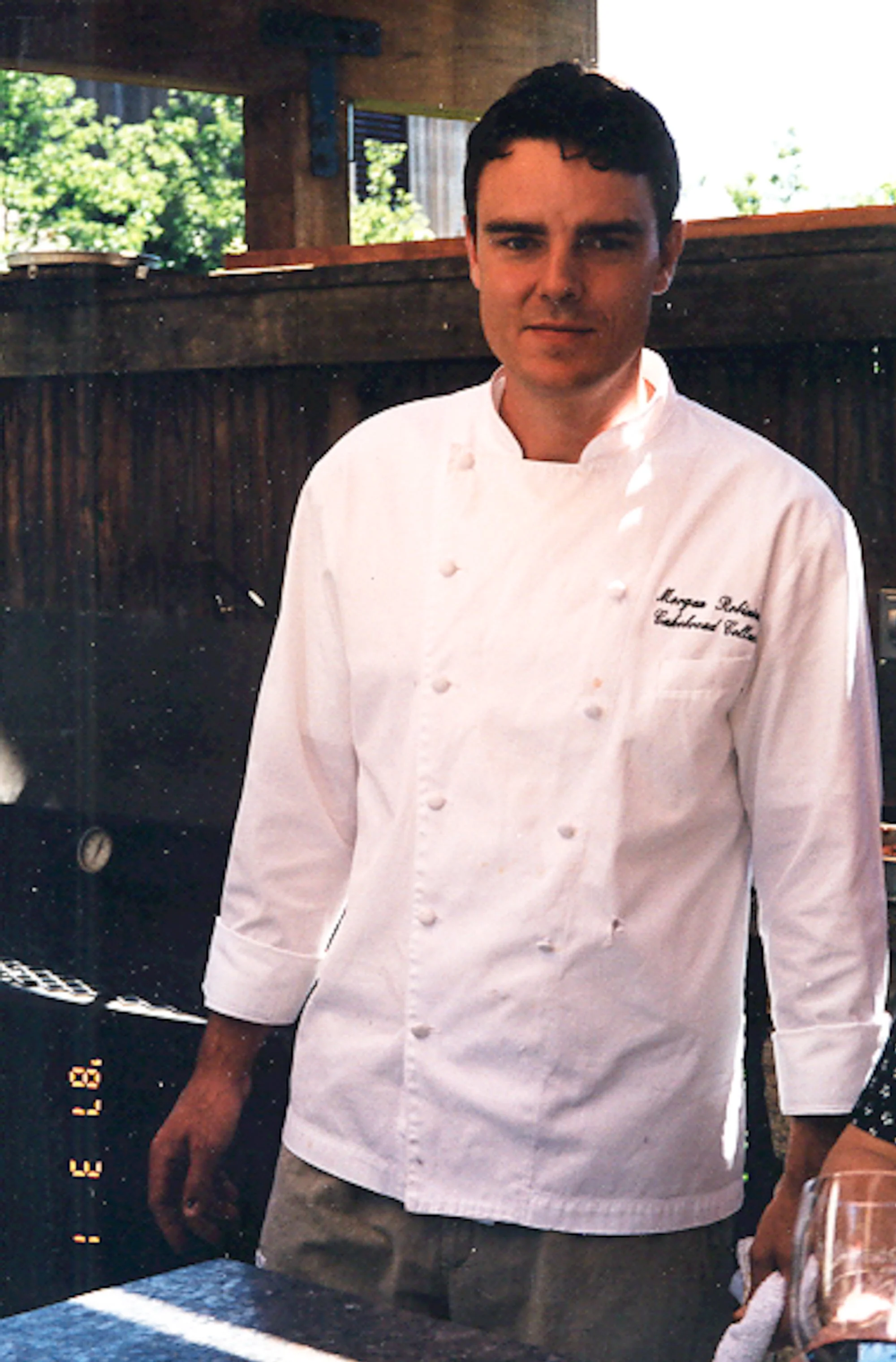 A male chef in a white chef coat standing near a grill or barbecue, with a wooden structure and green trees in the background.