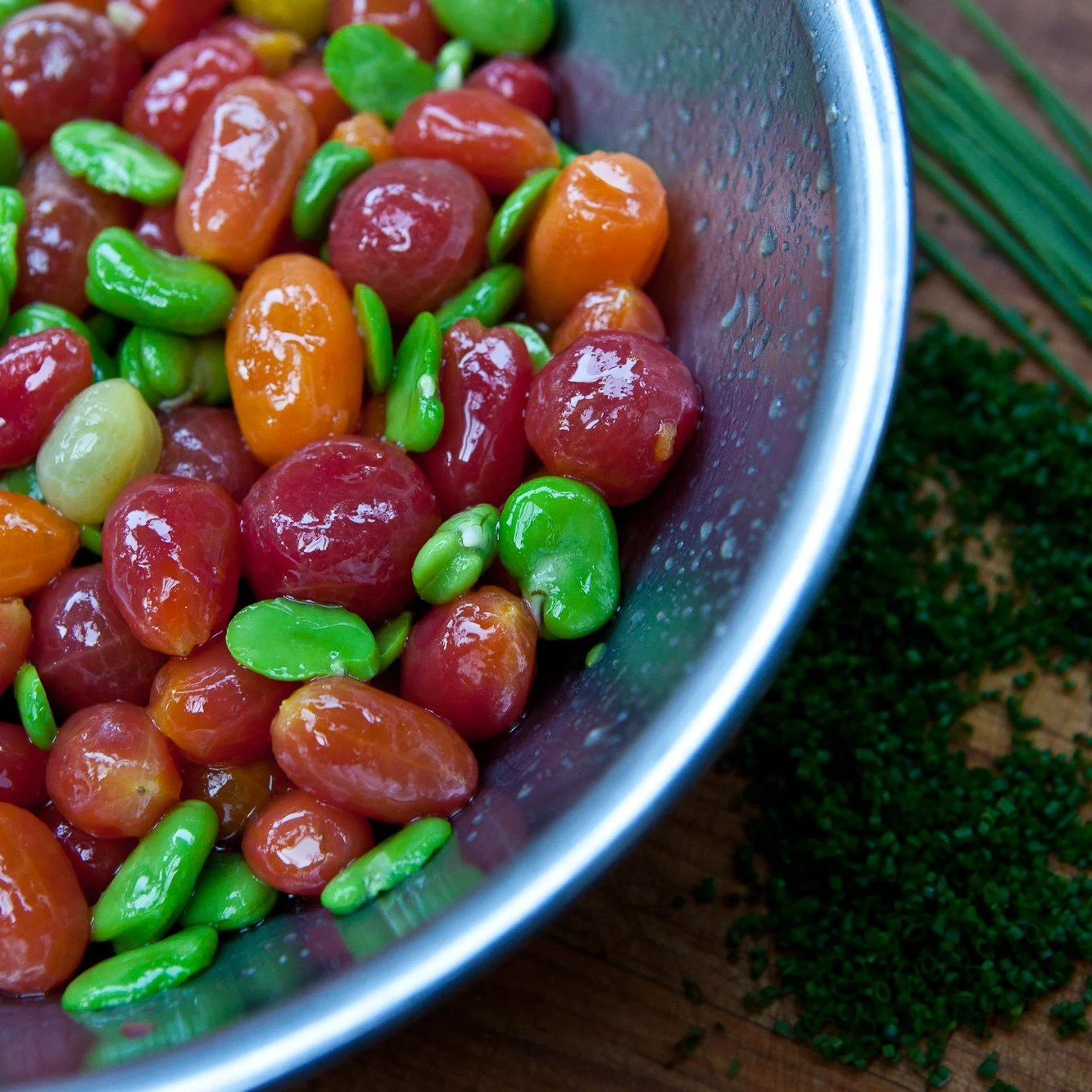 Close-up of a bowl filled with pickled cherries and green beans, with chopped herbs on a wooden surface.