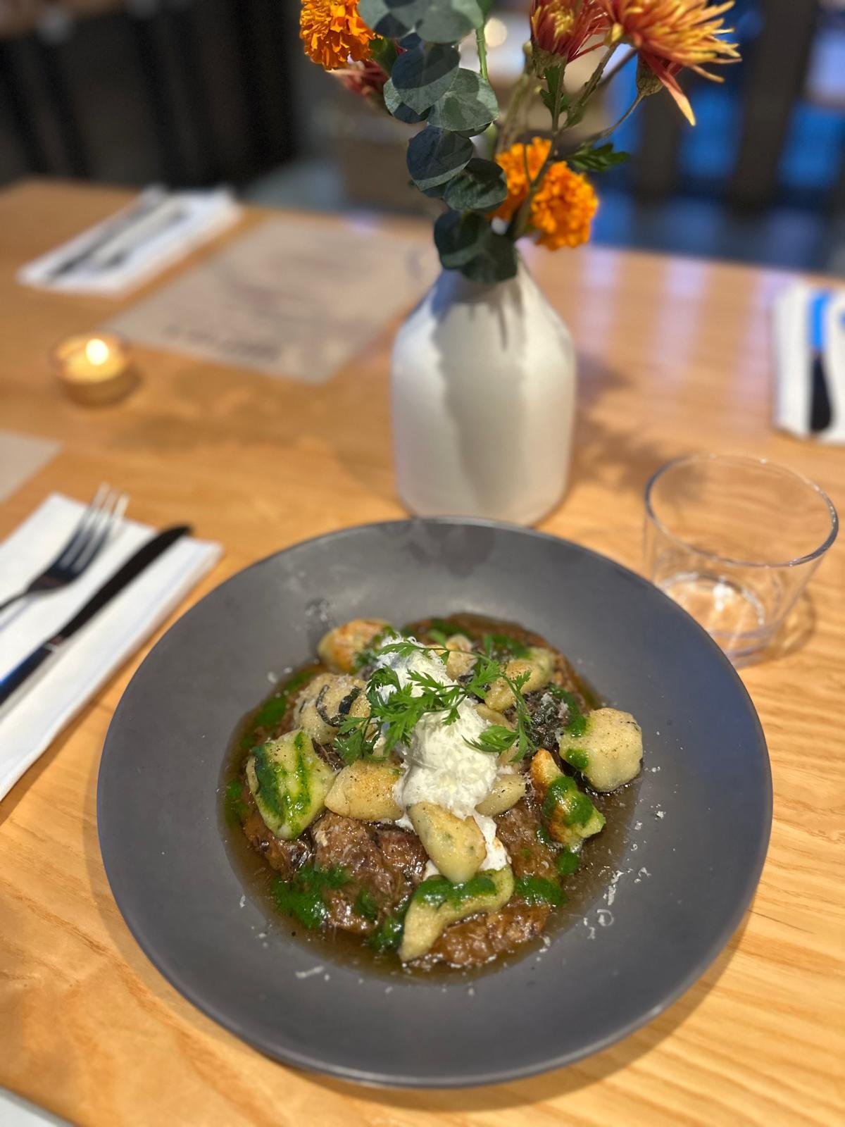 A plate of gnocchi with green sauce, topped with ricotta cheese and garnished with herbs, on a wooden table at a restaurant. In the background, there is a white vase with orange and yellow flowers.