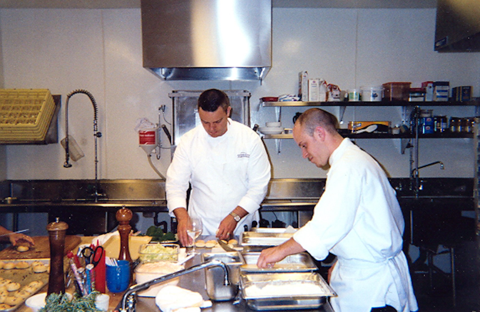 Two chefs in white uniforms working in a commercial kitchen, preparing food on a countertop with trays, ingredients, and kitchen tools visible.