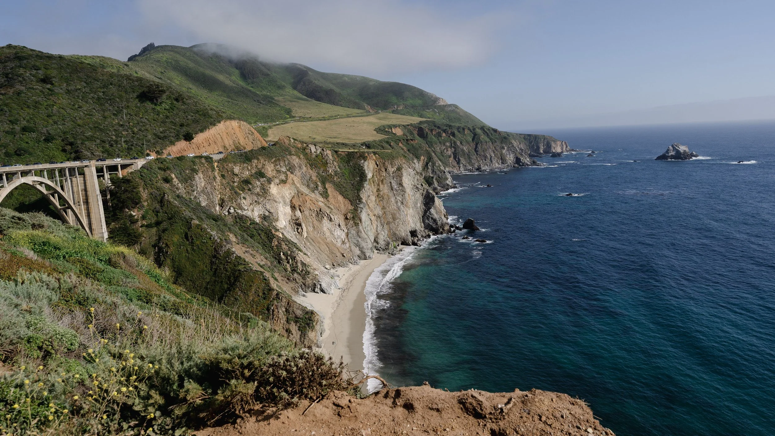 Coastal cliffs with a highway on top, overlooking a sandy beach and the ocean with rocks in the water.