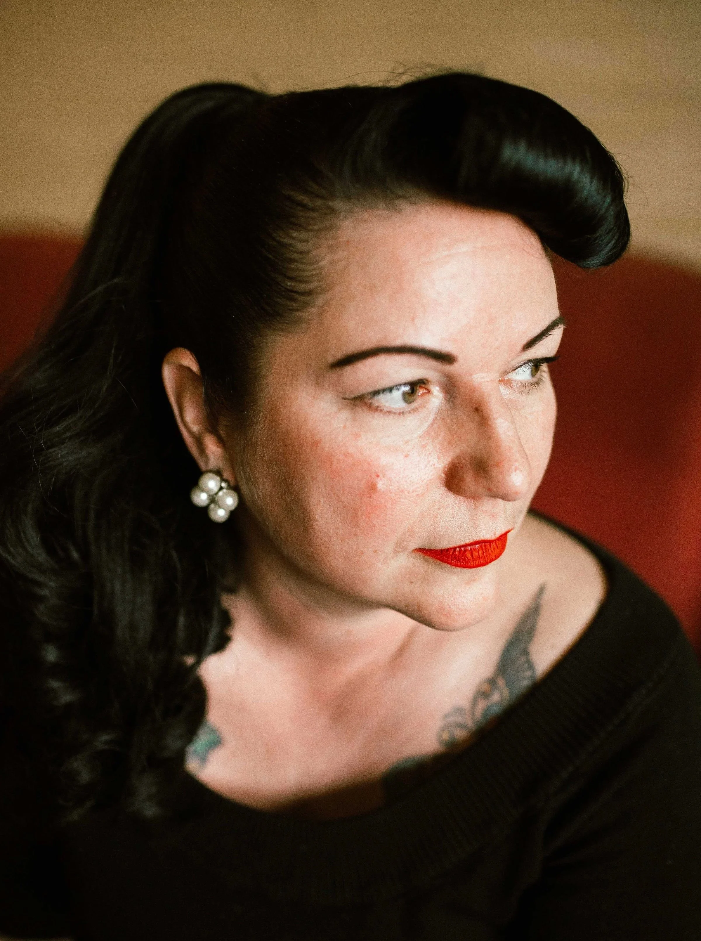 Close-up of a woman with black hair, wearing red lipstick, pearl earrings, and a black top, sitting on a red chair.