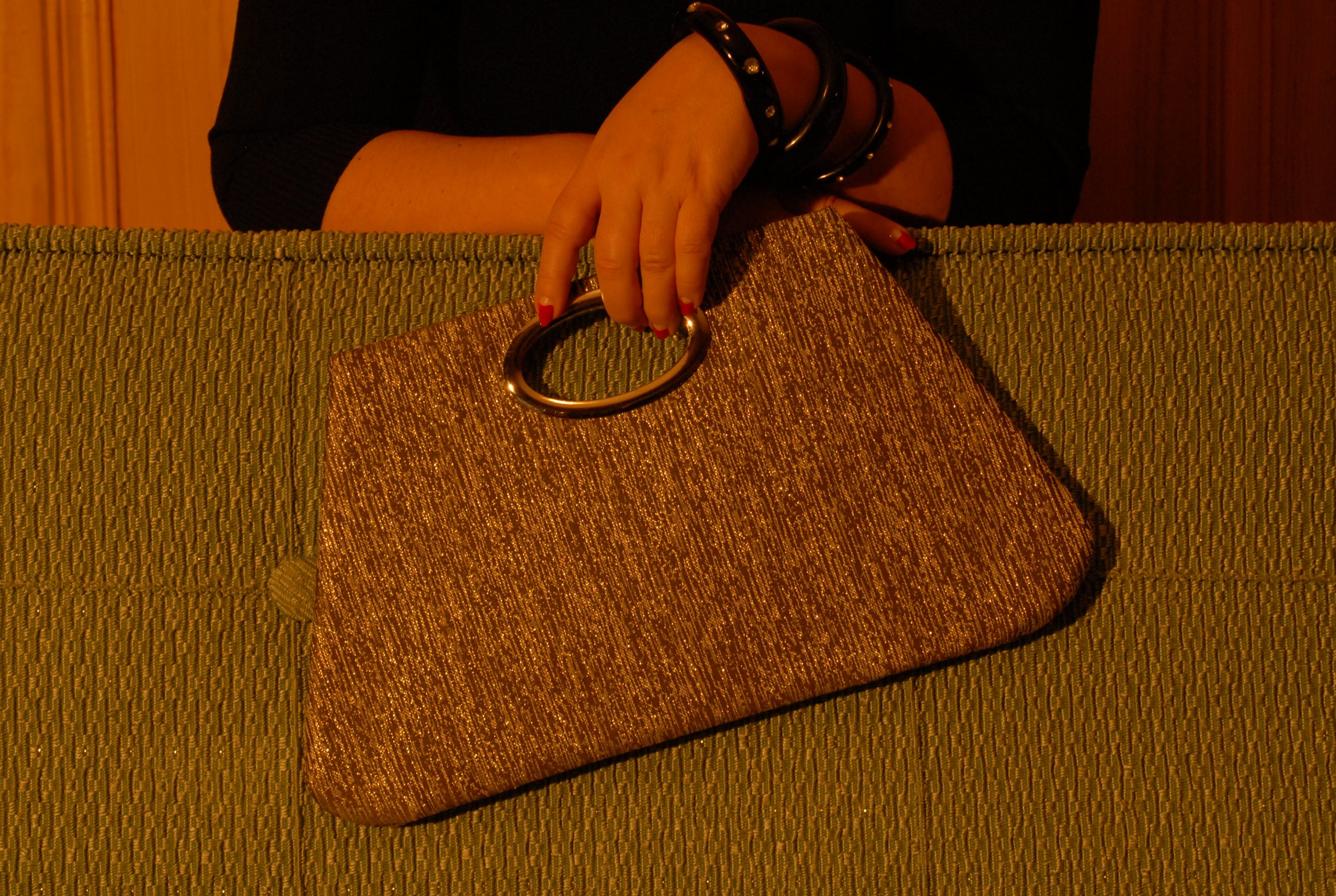 A person with red nail polish and black bracelets is holding a sparkling, textured gold clutch purse with circular handles, resting on a green upholstered surface.