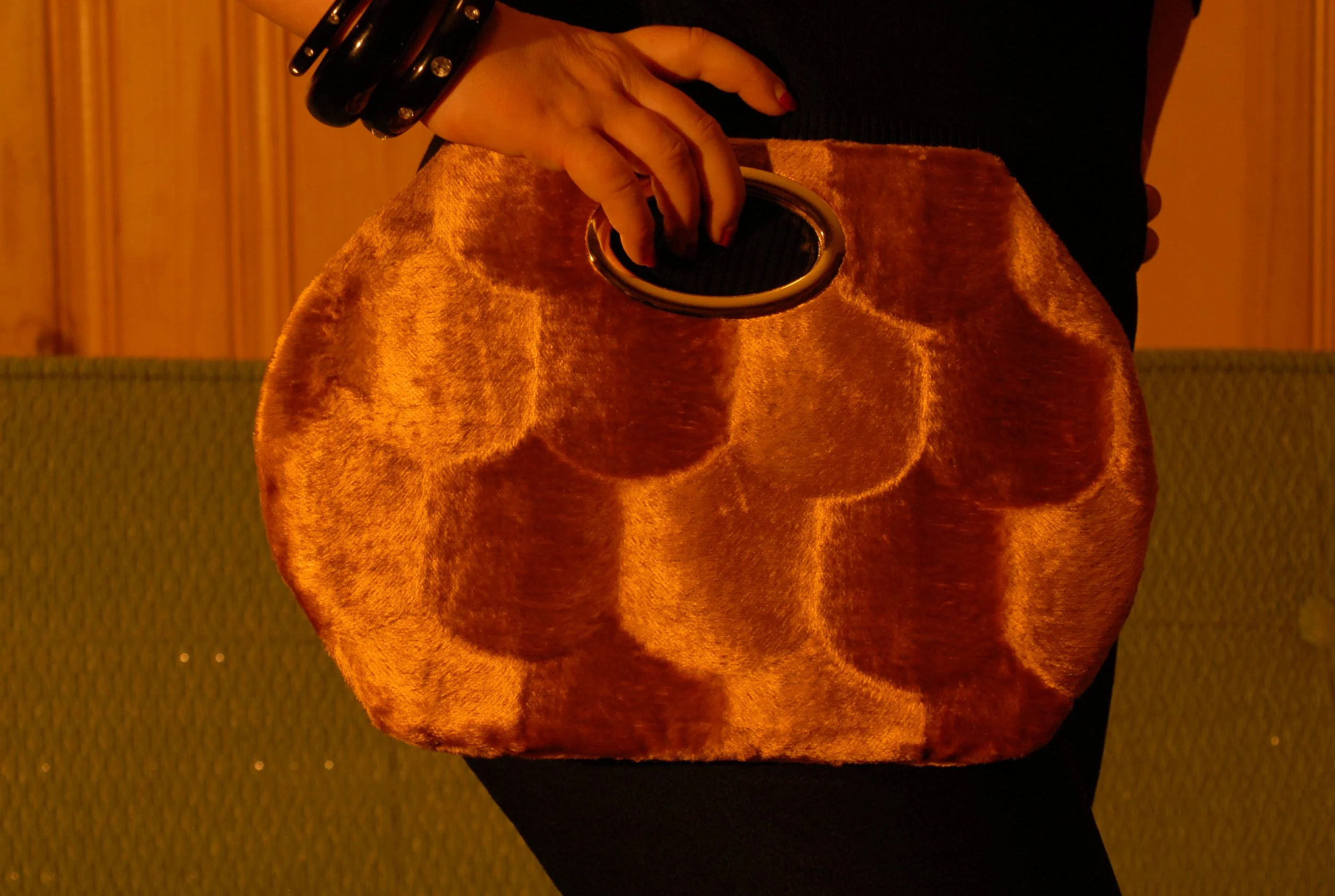 Close-up of a hand holding a brown velvet clutch purse with a circular opening and a black hand with bracelets against a wooden wall background.