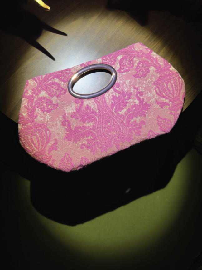 Pink and beige patterned clutch with a round metal handle on top, placed on a black covering, next to a wooden table.