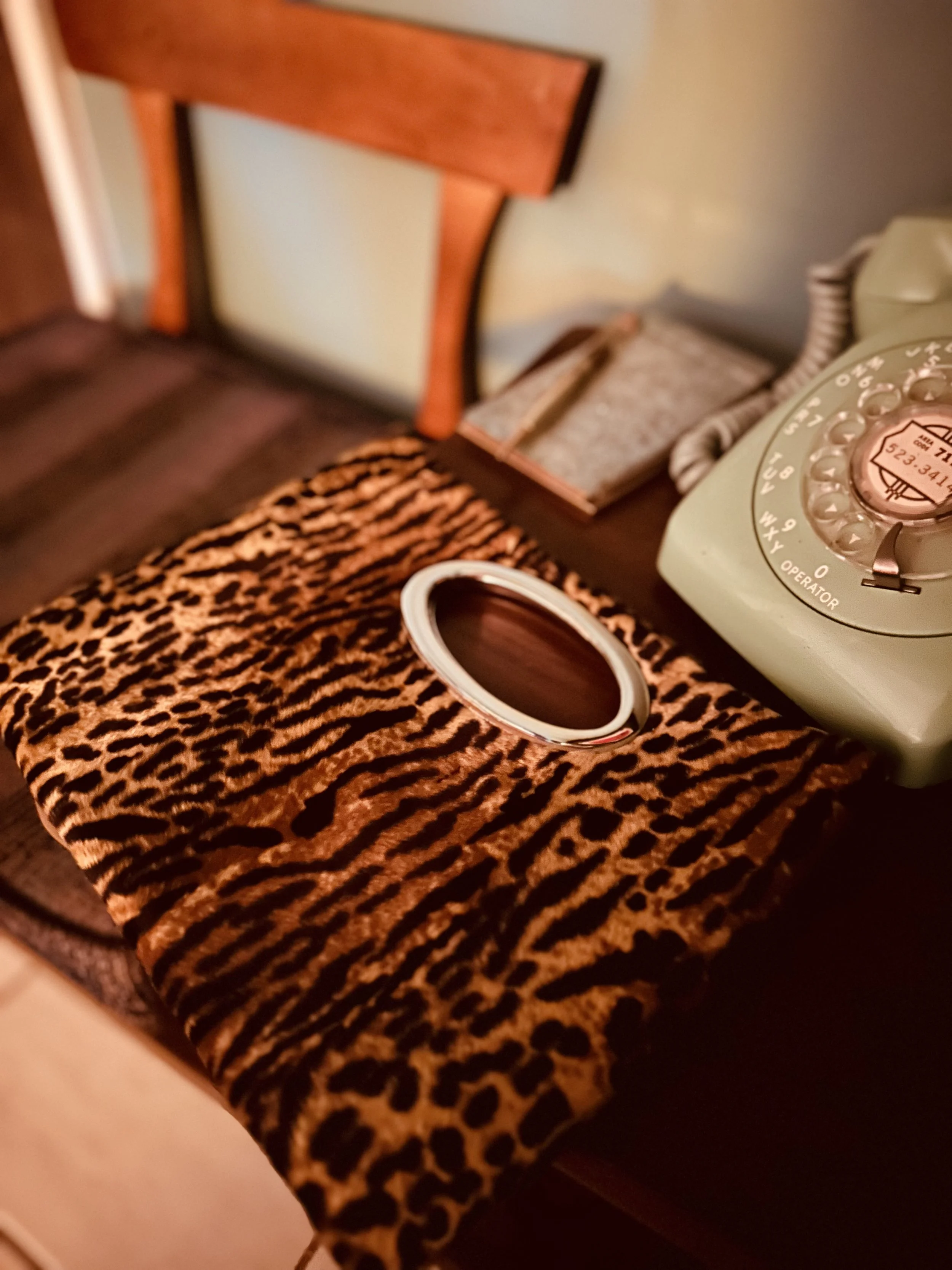 A vintage rotary dial telephone on a wooden table with a leopard print handbag and a wooden chair in the background.