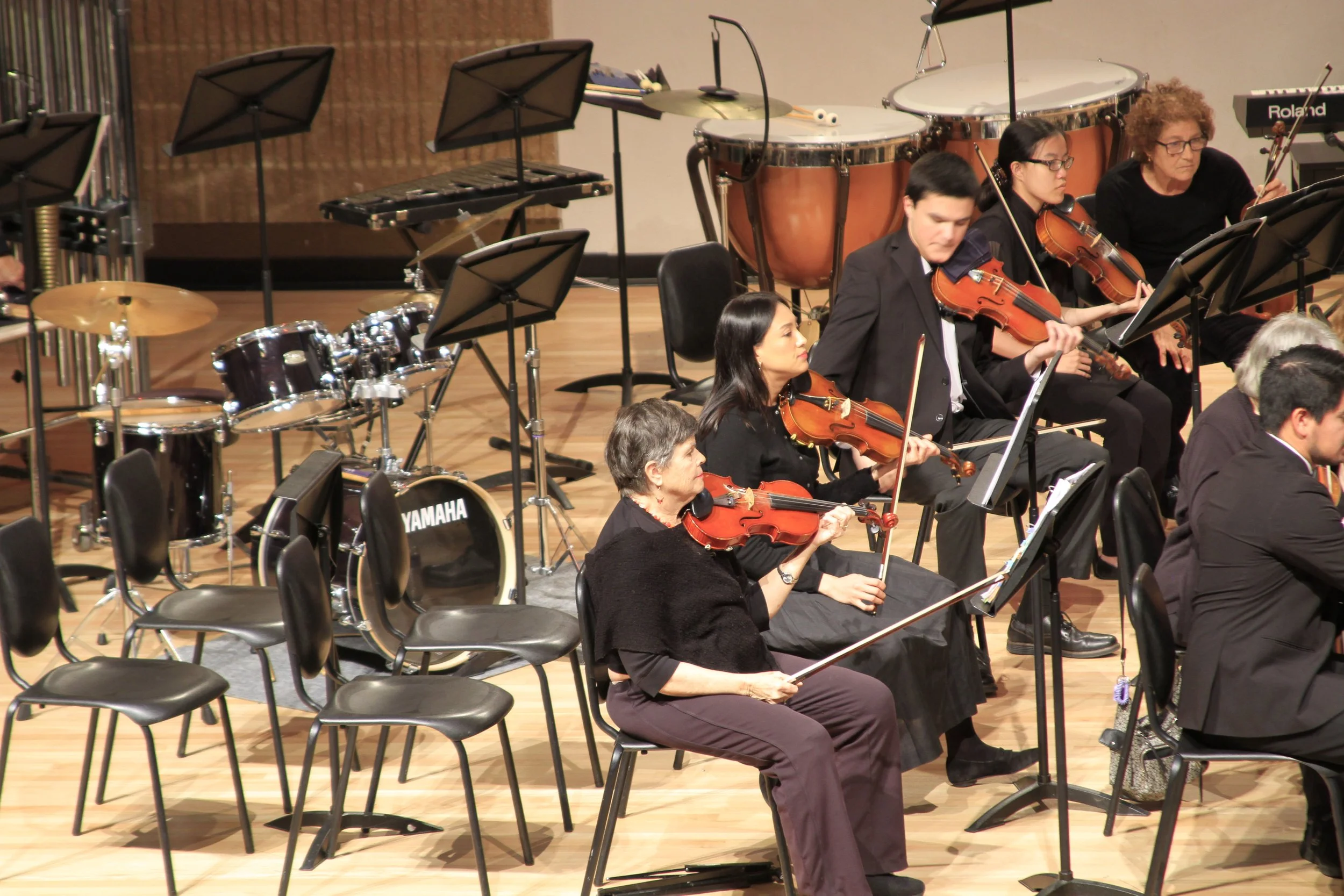 Orchestra members playing violins and other instruments, with drums and percussion in the background, on a wooden stage.