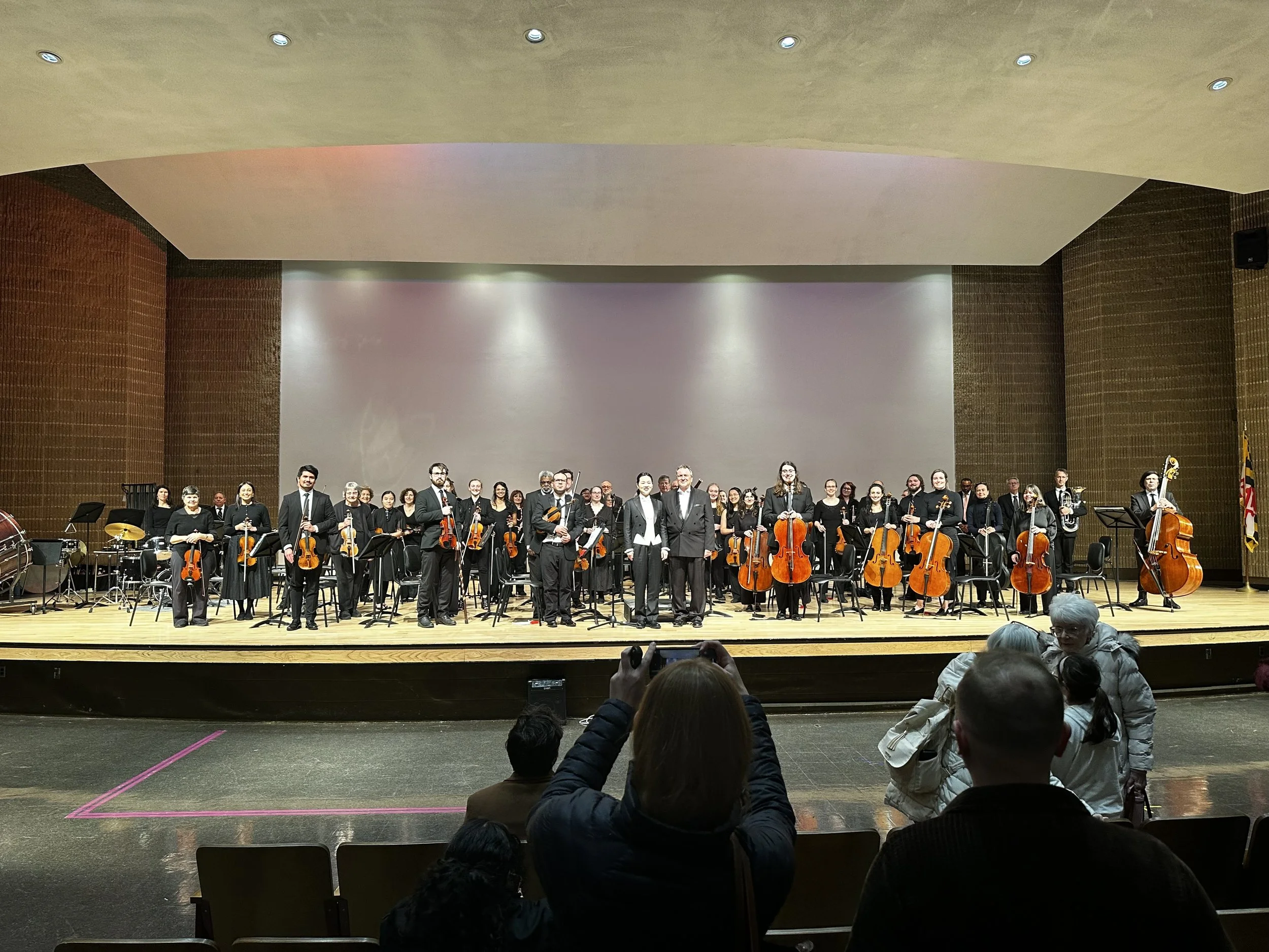 An orchestral ensemble on stage after a performance with members in formal black attire, including Swiftly playing violins, cellos, and other instruments, with binoculars and percussion instruments visible, in a concert hall with audience members in foreground taking photos.
