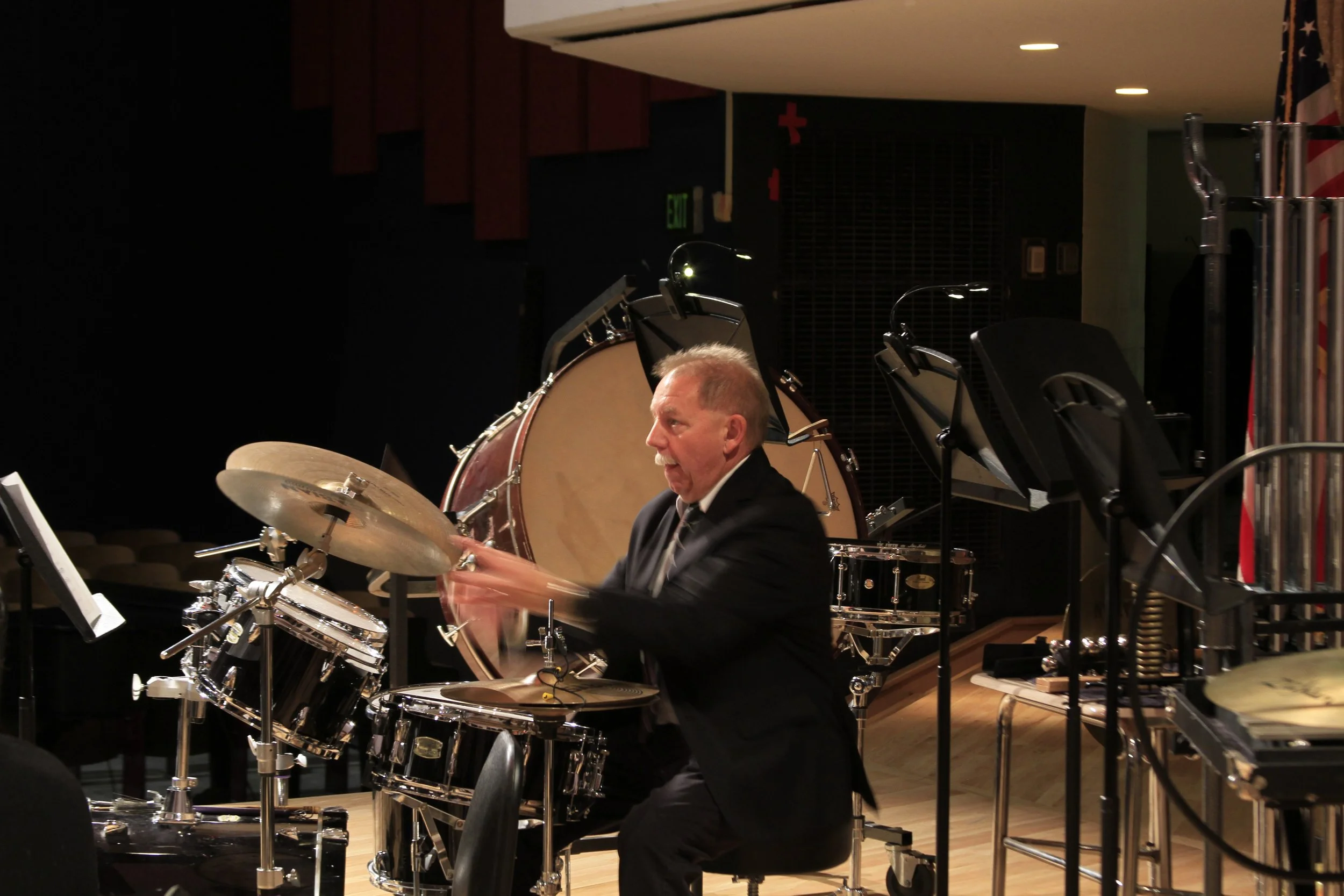 A man in a black suit playing drums on stage in a dark room with musical equipment and a large bass drum in the background.