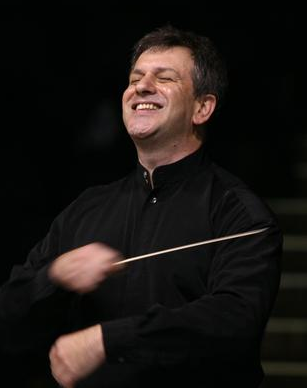 A man conducting an orchestra, smiling, holding a baton.