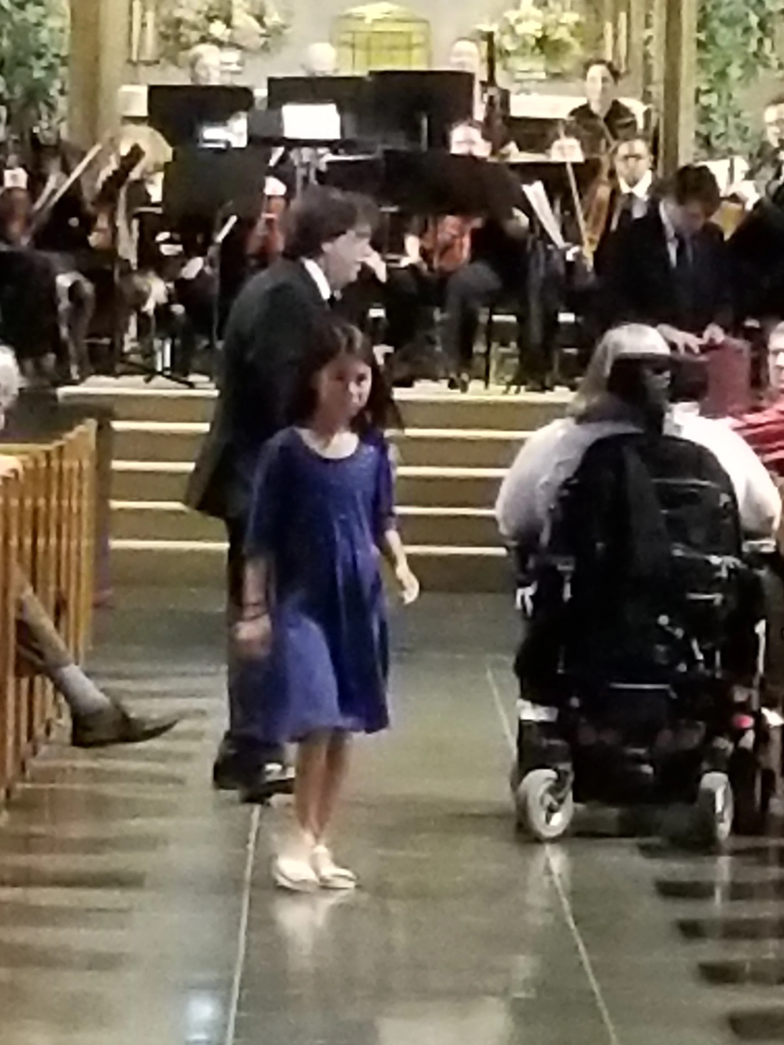 A girl in a blue dress walking in front of a crowded orchestra in a concert hall.