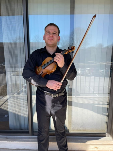 A man standing outside holding a violin and bow, dressed in black, with a glass window and building in the background.