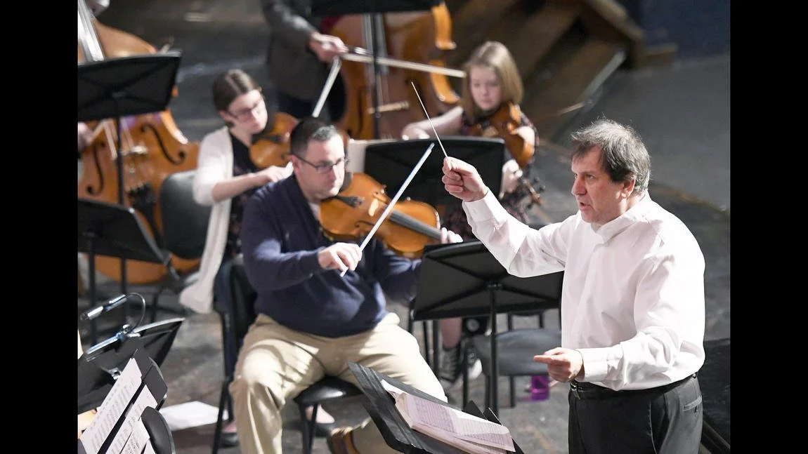 Conductor leading an orchestra with violinists in the background.