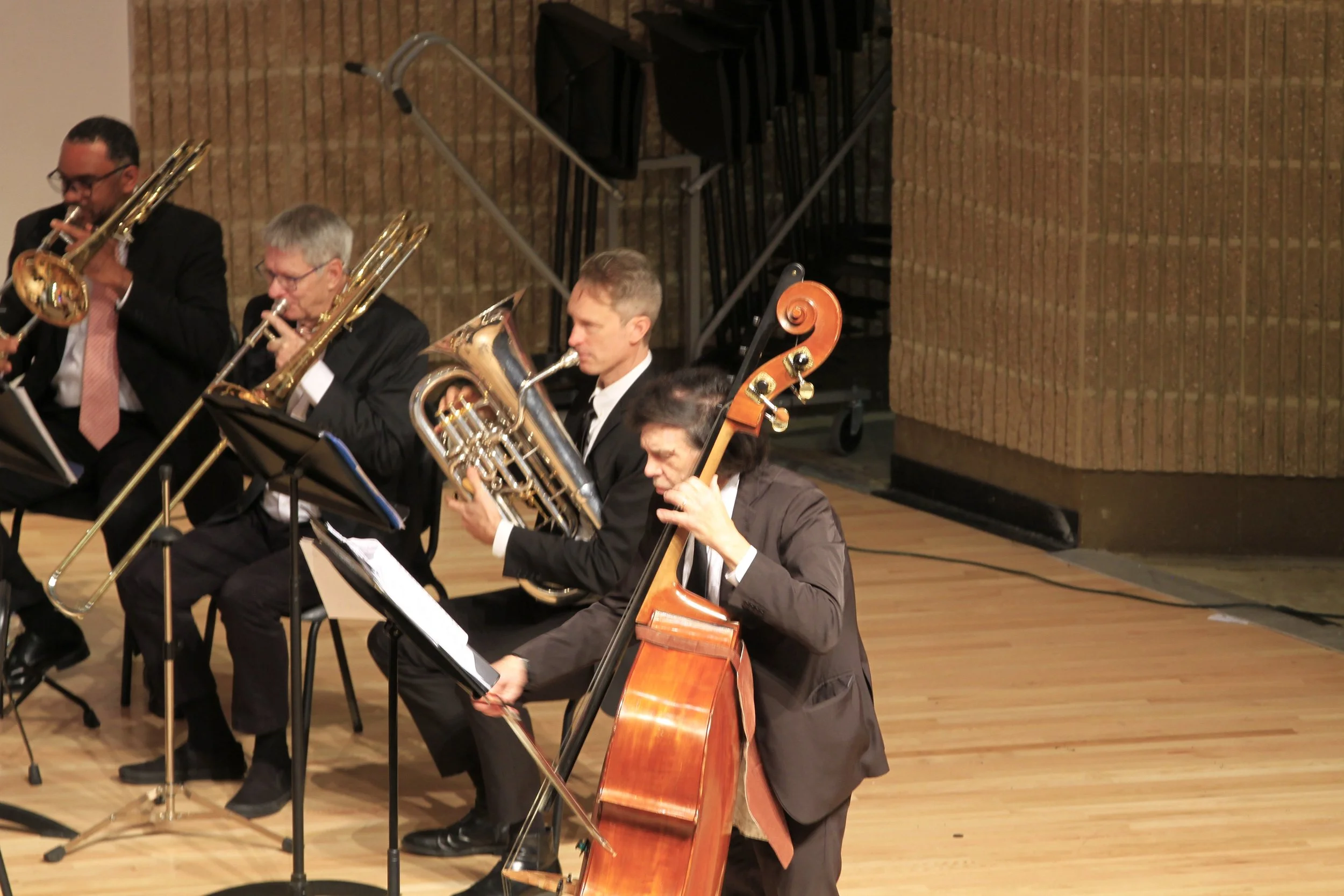 An orchestra of musicians performing on stage, playing various instruments including trombones, French horn, and double bass, with music stands and sheet music in front of them.