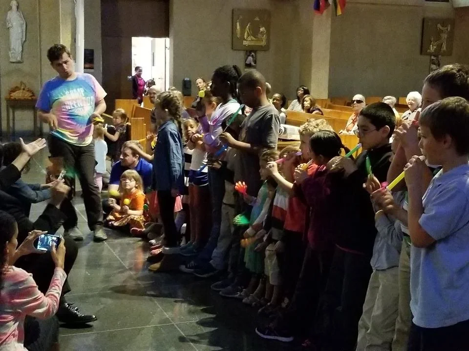 Group of children and adults gathered in a church, some playing flutes, with some taking photos, and others sitting in pews, during a celebration or event.