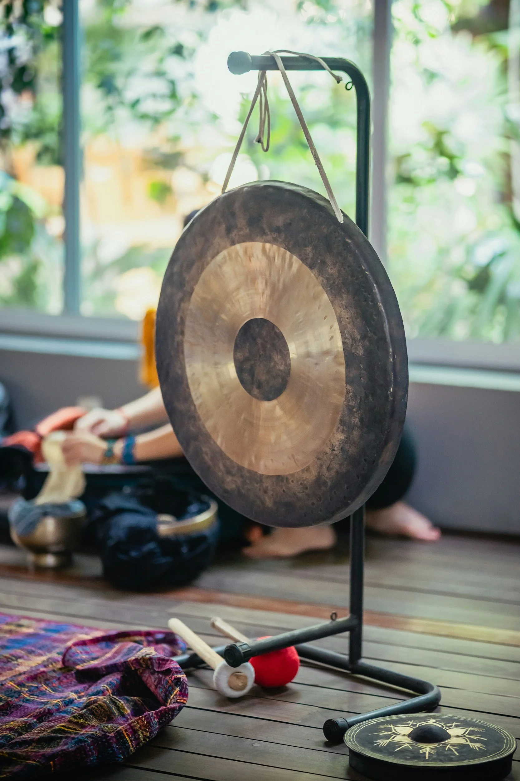 Crystal singing bowl with a metal stand, surrounded by colorful fabric, a mallet, a red ball, a drum, and various musical instruments on a wooden floor with a window and greenery in the background.