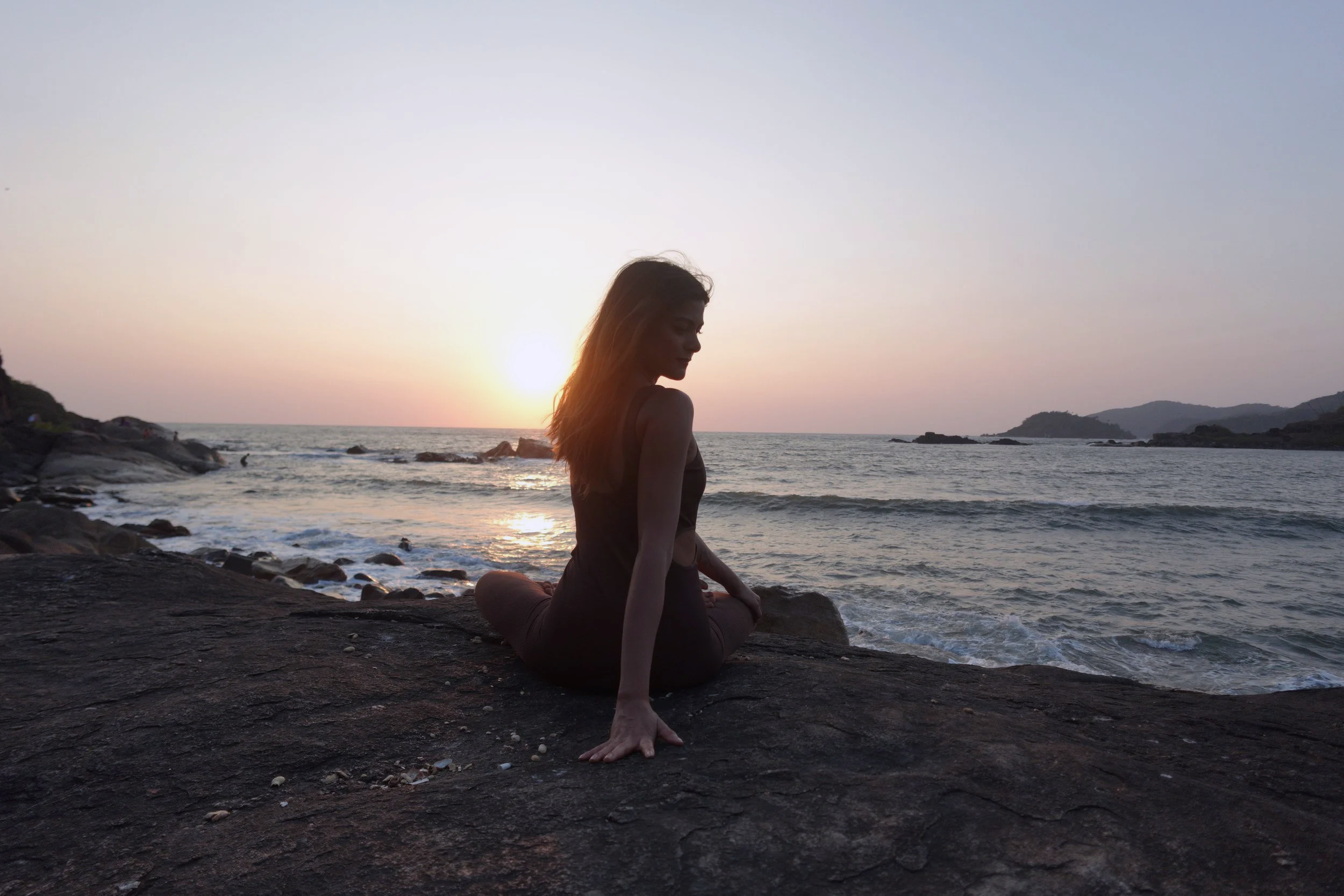 A woman practicing yoga on rocks by the ocean during sunset.
