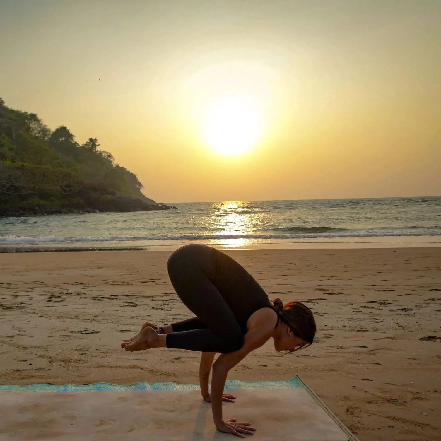 A woman practicing yoga on a beach during sunset, in a crow pose with hands on the ground, knees resting on her arms, facing the ocean with trees on a hill in the background.