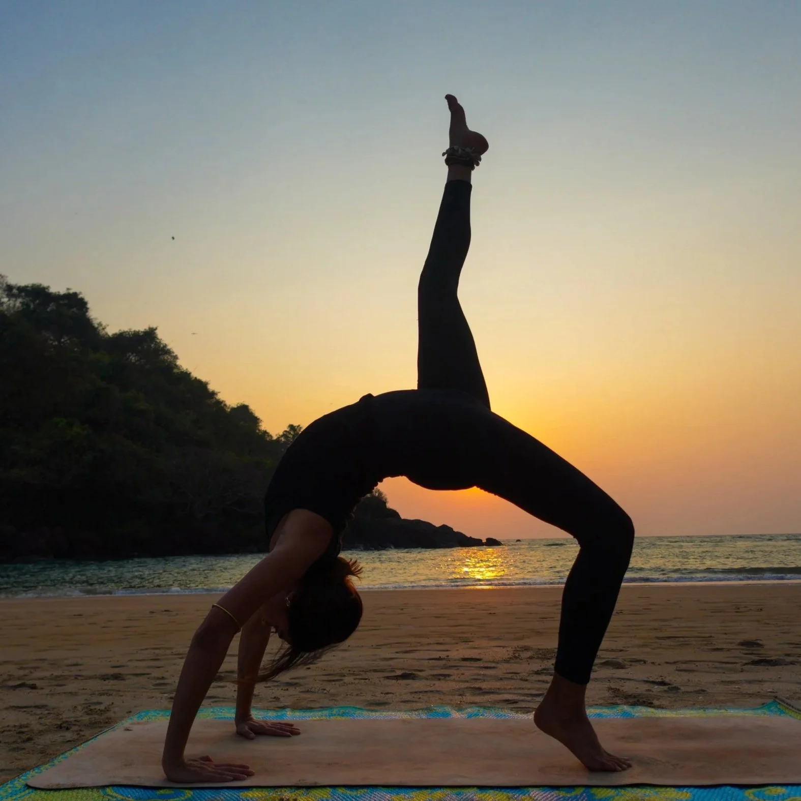A person doing a yoga pose on a beach at sunset, with the sky in shades of orange and blue and water in the background.
