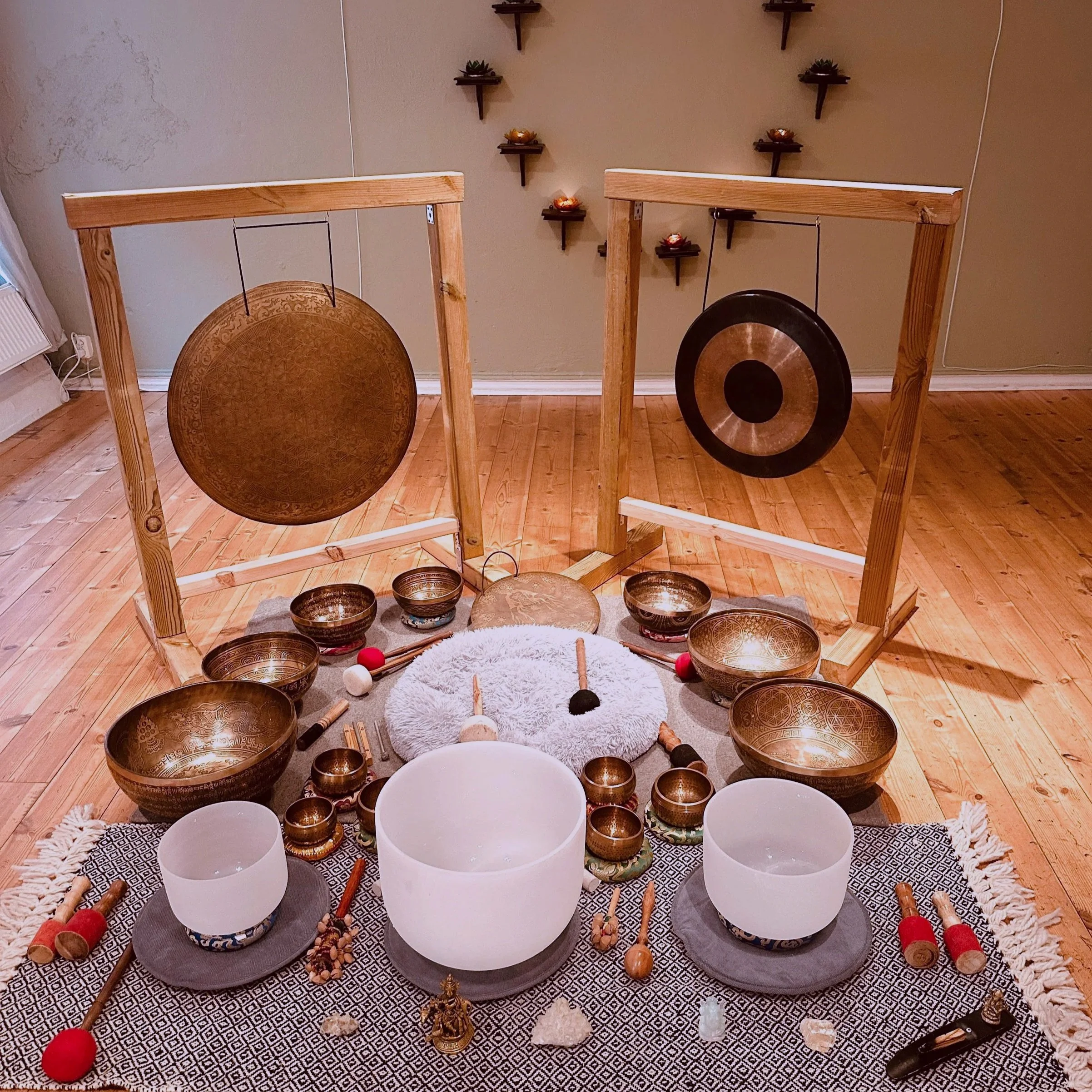 A set of metal singing bowls of various sizes placed on a patterned cloth. Behind the bowls, a wooden frame holds three white bowls, and in front of it, several small brass and decorative bowls are arranged. Items like mallets and small figurines surround the bowls, with a soft fleece rug underneath the setup and a gray and white geometric patterned cloth.