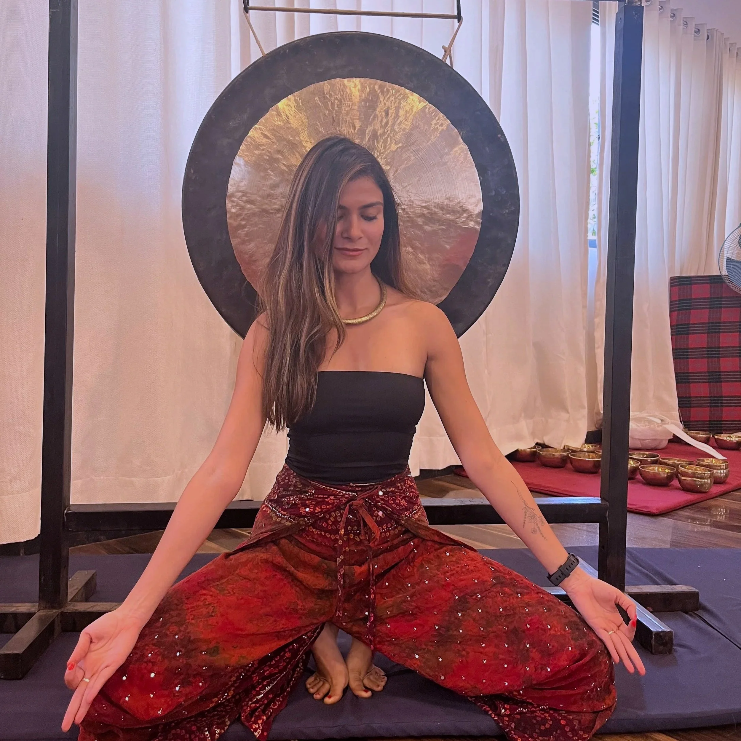 A woman with long brown hair sitting cross-legged in front of a large gong, with her eyes closed and peaceful expression. She is wearing a black tube top, a red patterned skirt, a gold necklace, and a black wristband. The background shows a white curtain, some bowls, and a plaid chair.