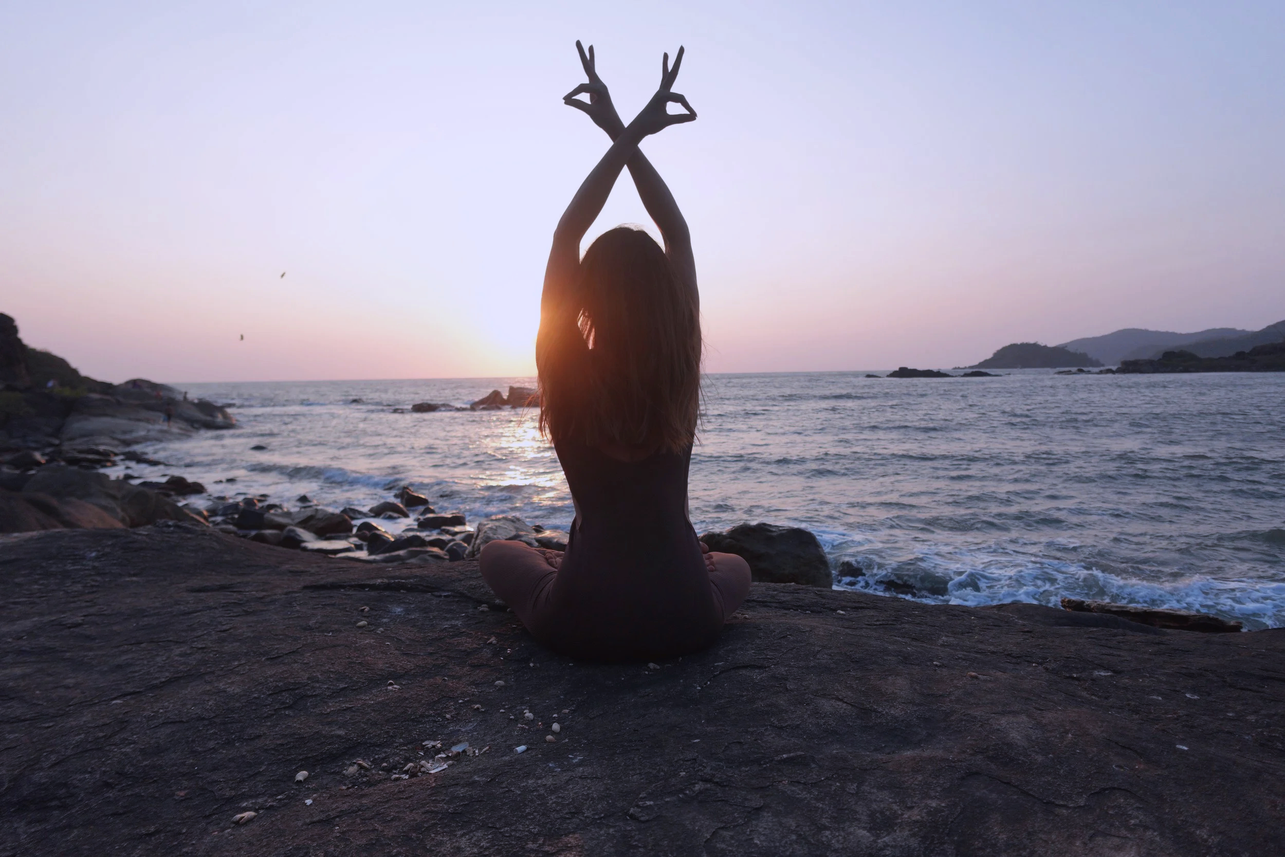 A person practicing yoga on a rocky beach at sunset, sitting cross-legged with arms raised and fingers in a mudra, facing the ocean with mountains in the distance.