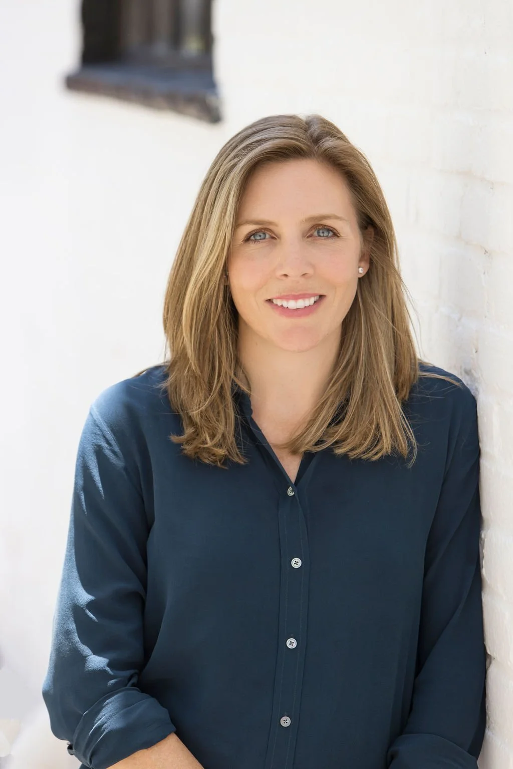 A woman with shoulder-length light brown hair, wearing a navy blue button-up shirt, smiling, leaning against a white brick wall.