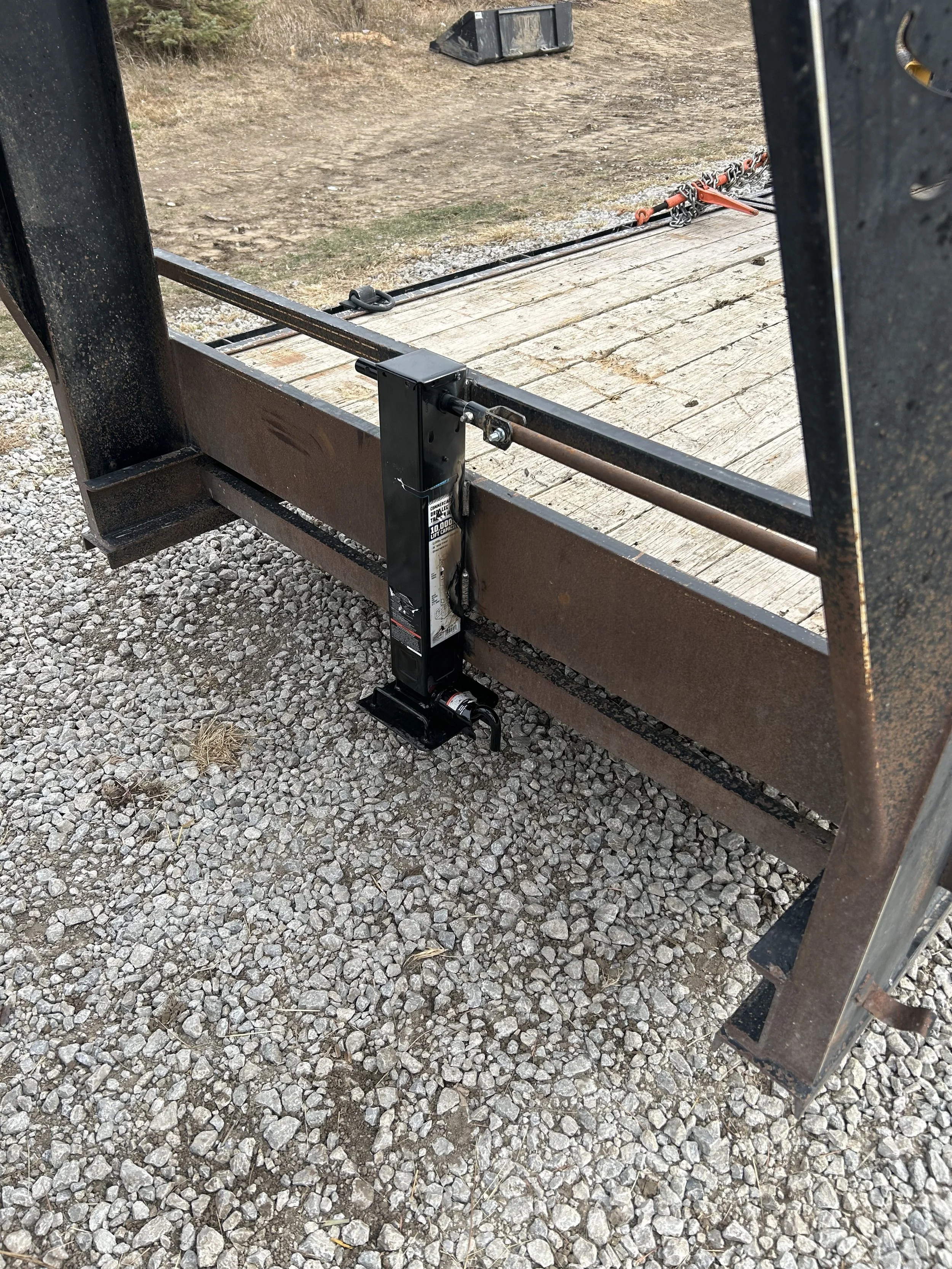 Close-up of a trailer hitch mechanism attached to a metal and wooden trailer, set on gravel ground with a black jack stand at the hitch.