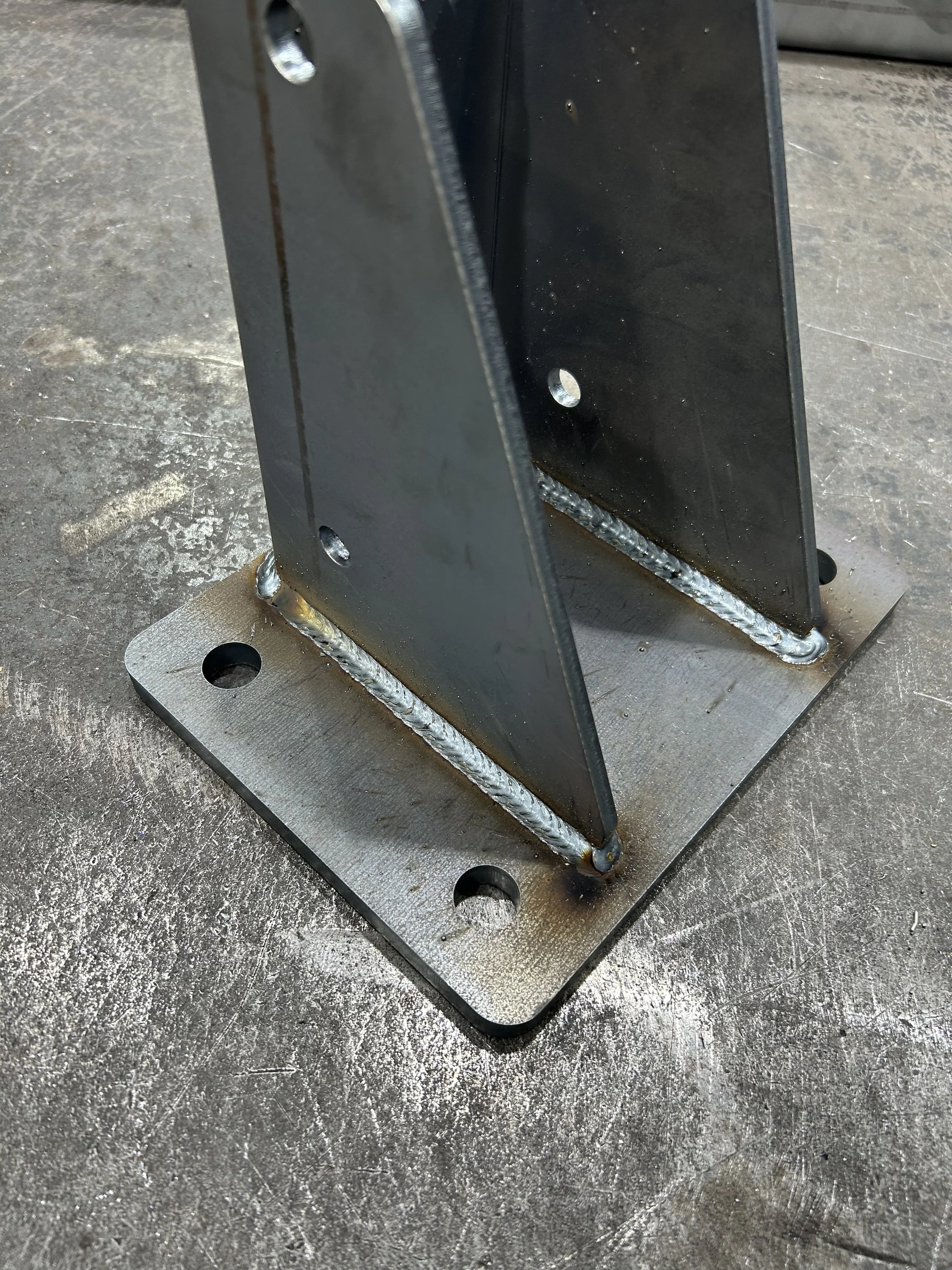 A welded metal base with two vertical metal plates attached at an angle, secured to a flat metal plate with four bolt holes.