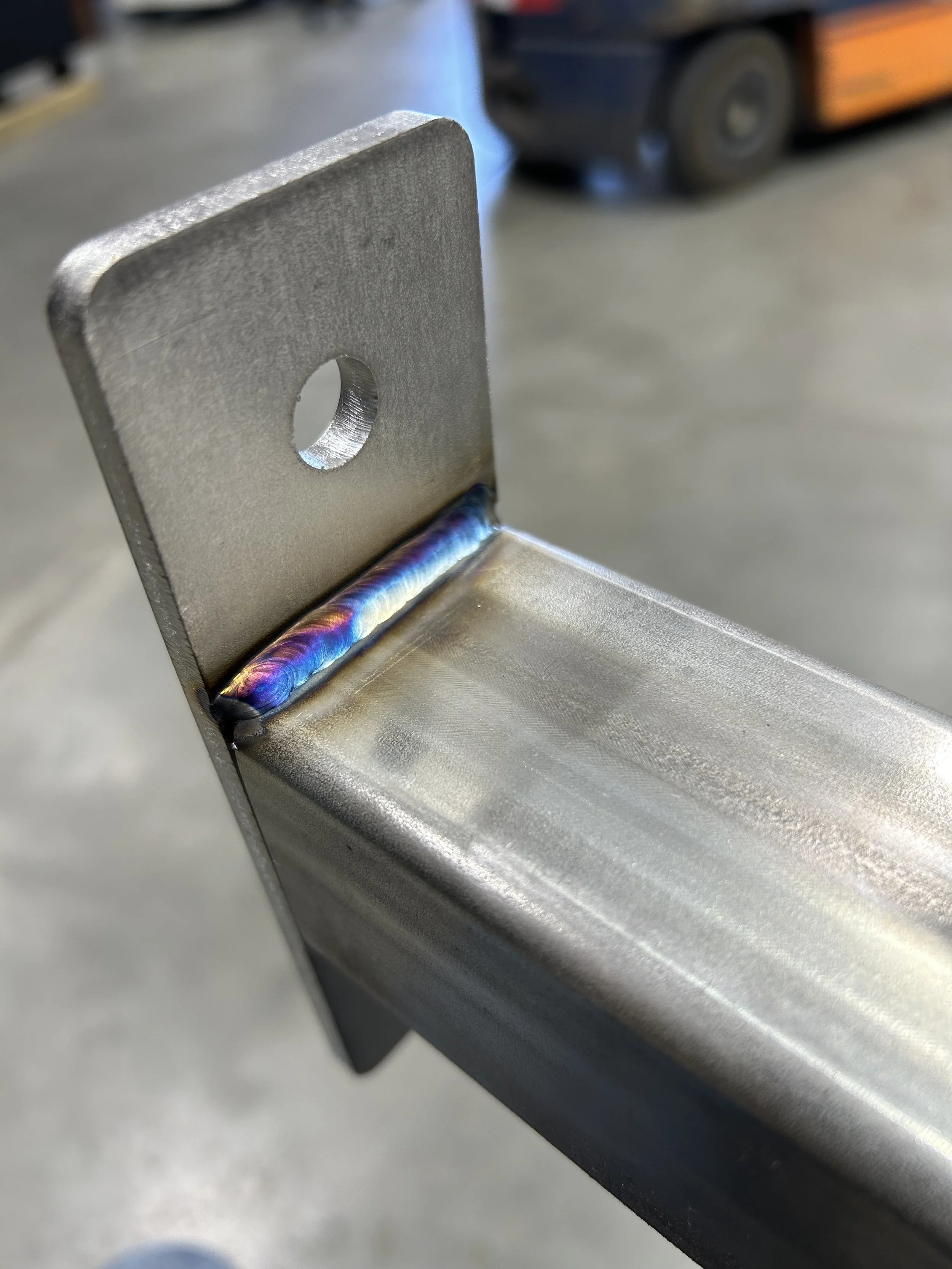 Close-up of a welded metal joint showing a colorful weld bead connecting a metal plate to a metal tube, with an industrial background.