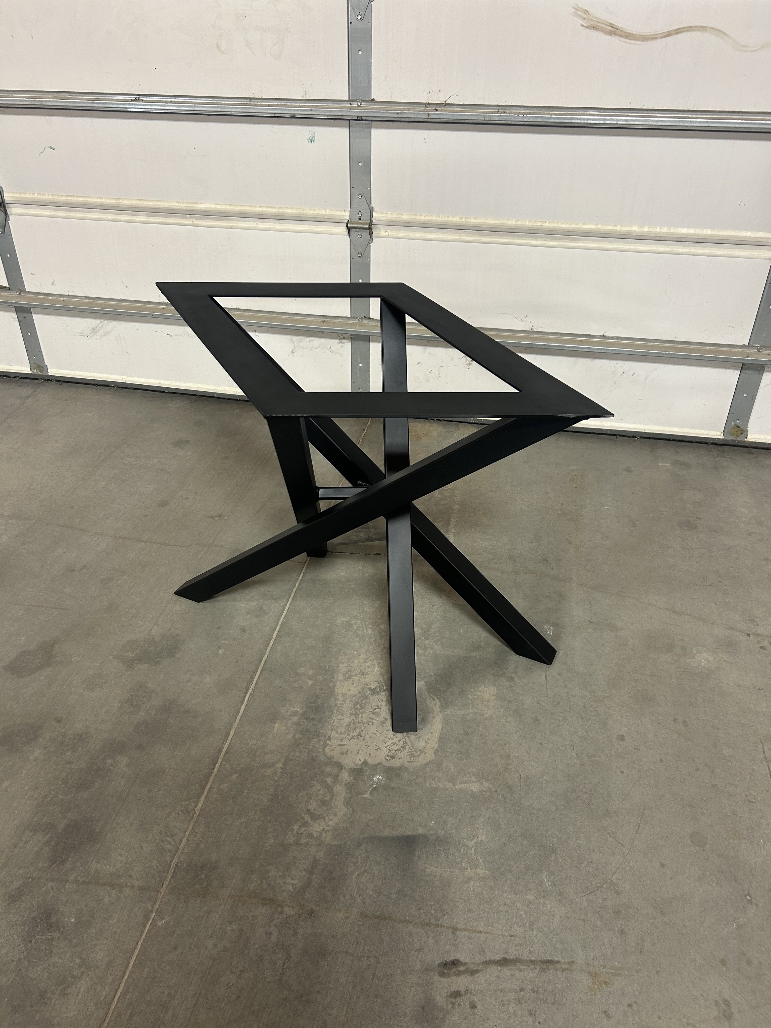 A black, minimalist table with an X-shaped base and a rectangular open frame on top, set against a white garage door.