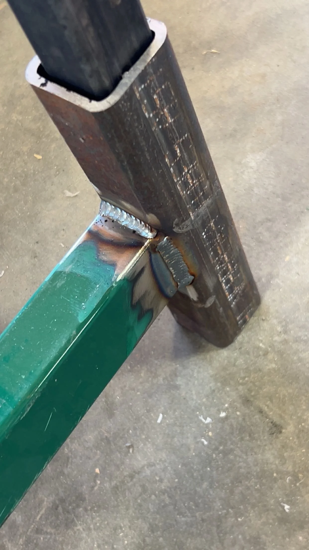 Close-up of a metal weld joint connecting a black steel tube to a painted green metal beam, with visible welding beads.