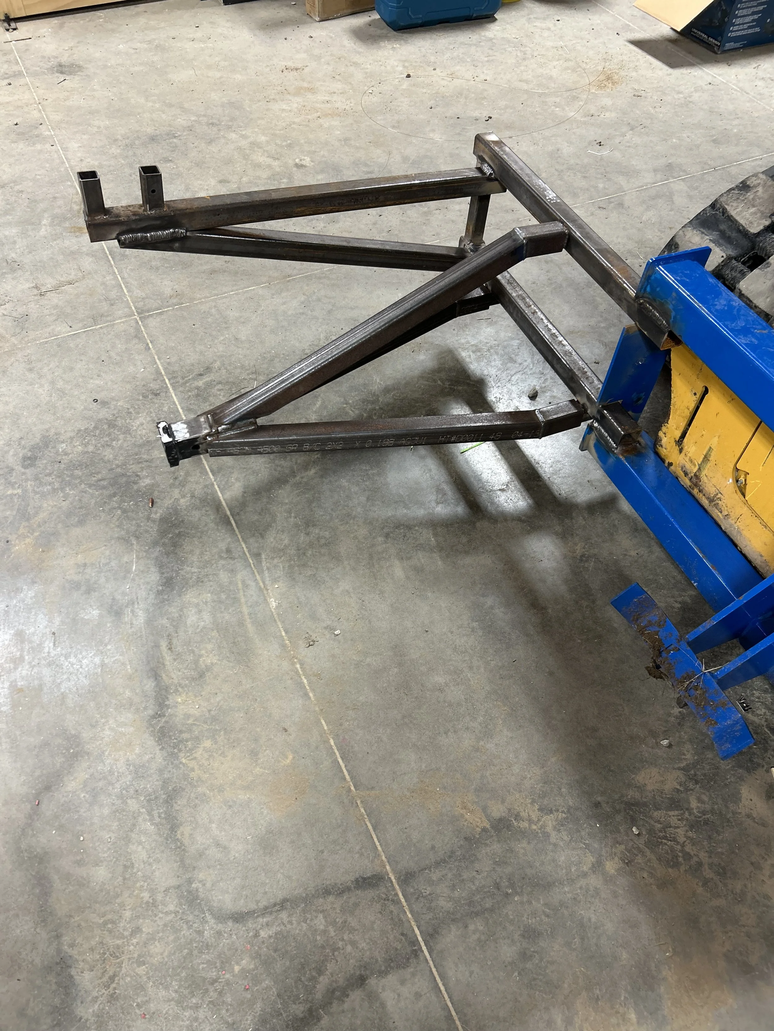 A metal frame in a workshop setting, attached to a blue and yellow workbench, with a concrete floor underneath.