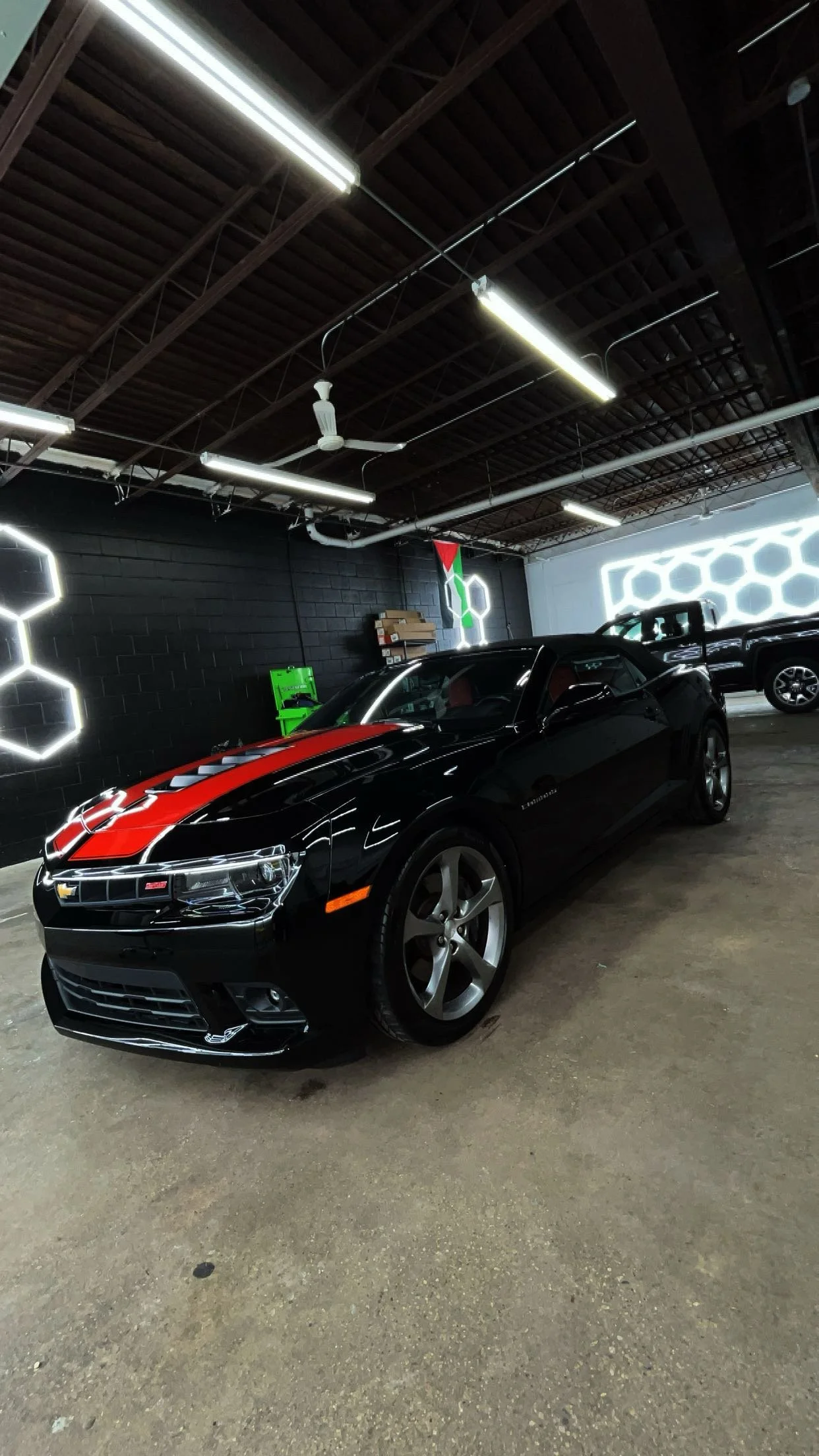 A black Chevrolet Camaro sports car with red racing stripes parked inside a modern showroom with geometric neon light decor on the walls.