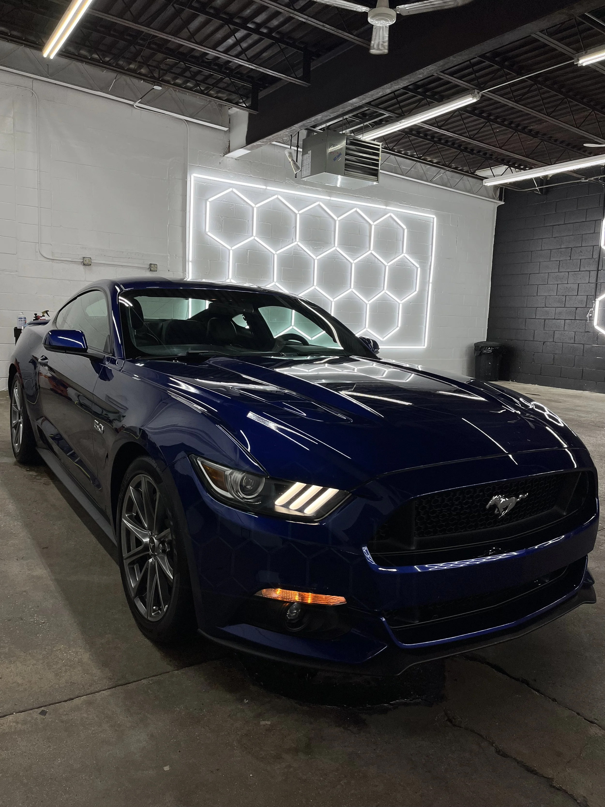 A blue Ford Mustang sports car parked inside a modern garage with white and black walls, illuminated by LED strip lights and decorated with a neon hexagon wall art.