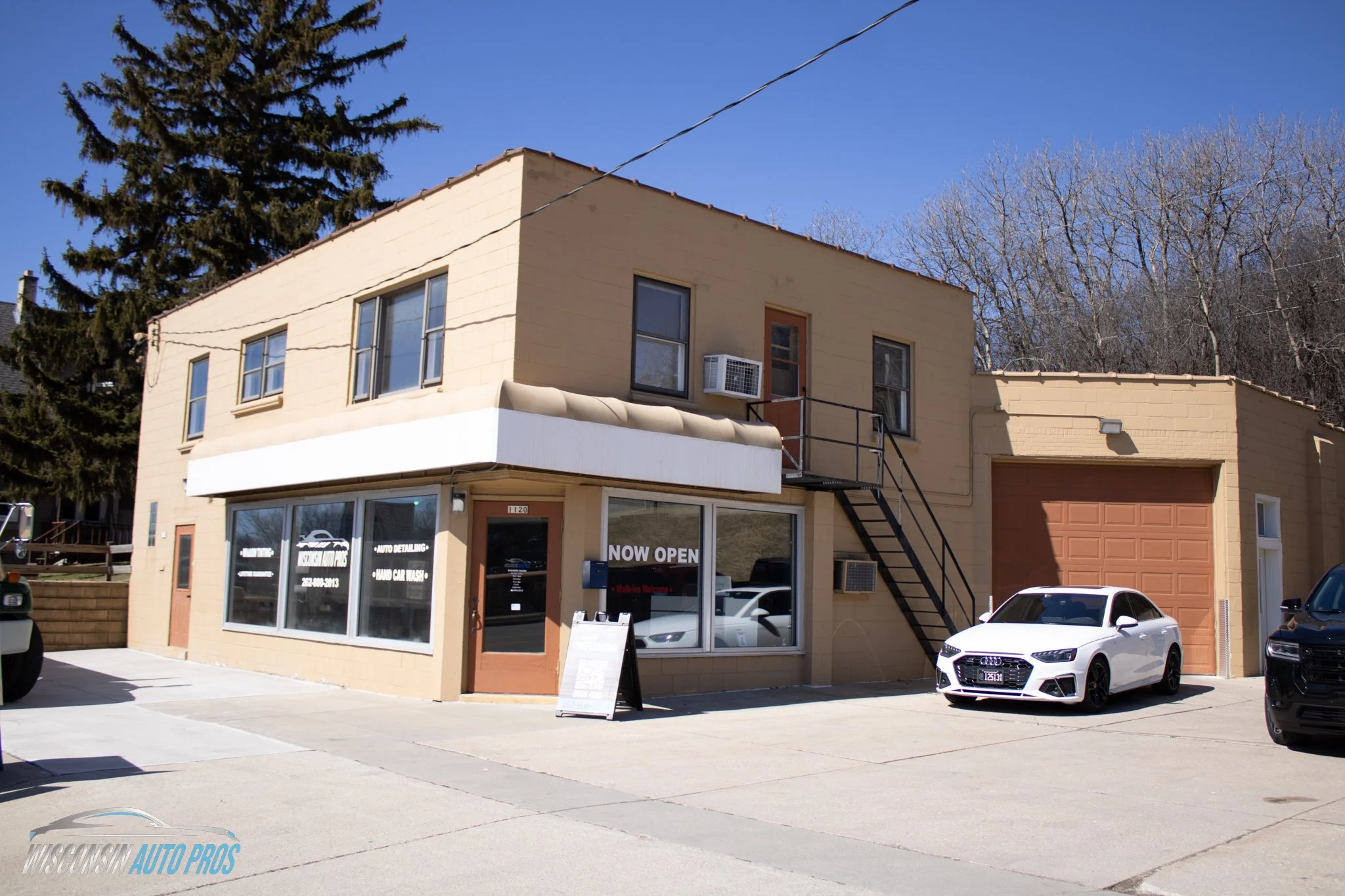 A two-story beige building with a car dealership on the ground floor, indicated by large windows and signs that read 'Now Open' and 'Auto Detailing, Hand Car Wash.' There are cars parked in front, including a white sedan closer to the building. A staircase leads to an upper door on the right side of the building. The sky is clear and blue, with some trees in the background.