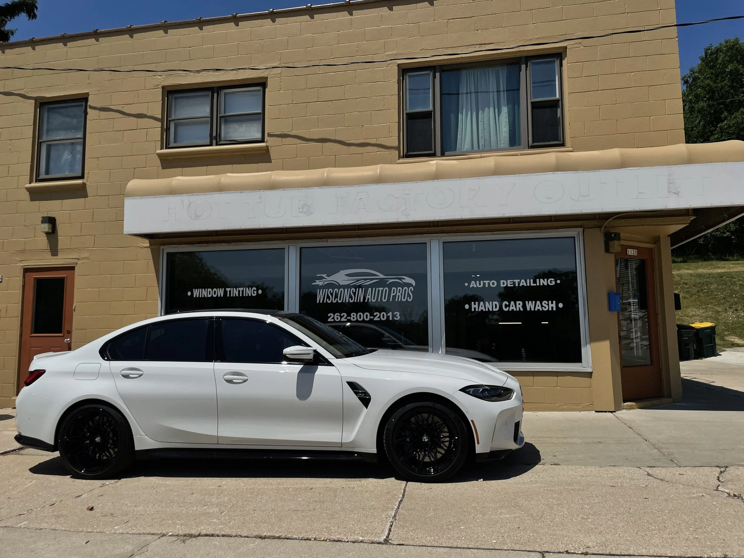 White sedan parked in front of a beige building with signage for Wisconsin Auto Pros, offering window tinting, auto detailing, and hand car wash services.