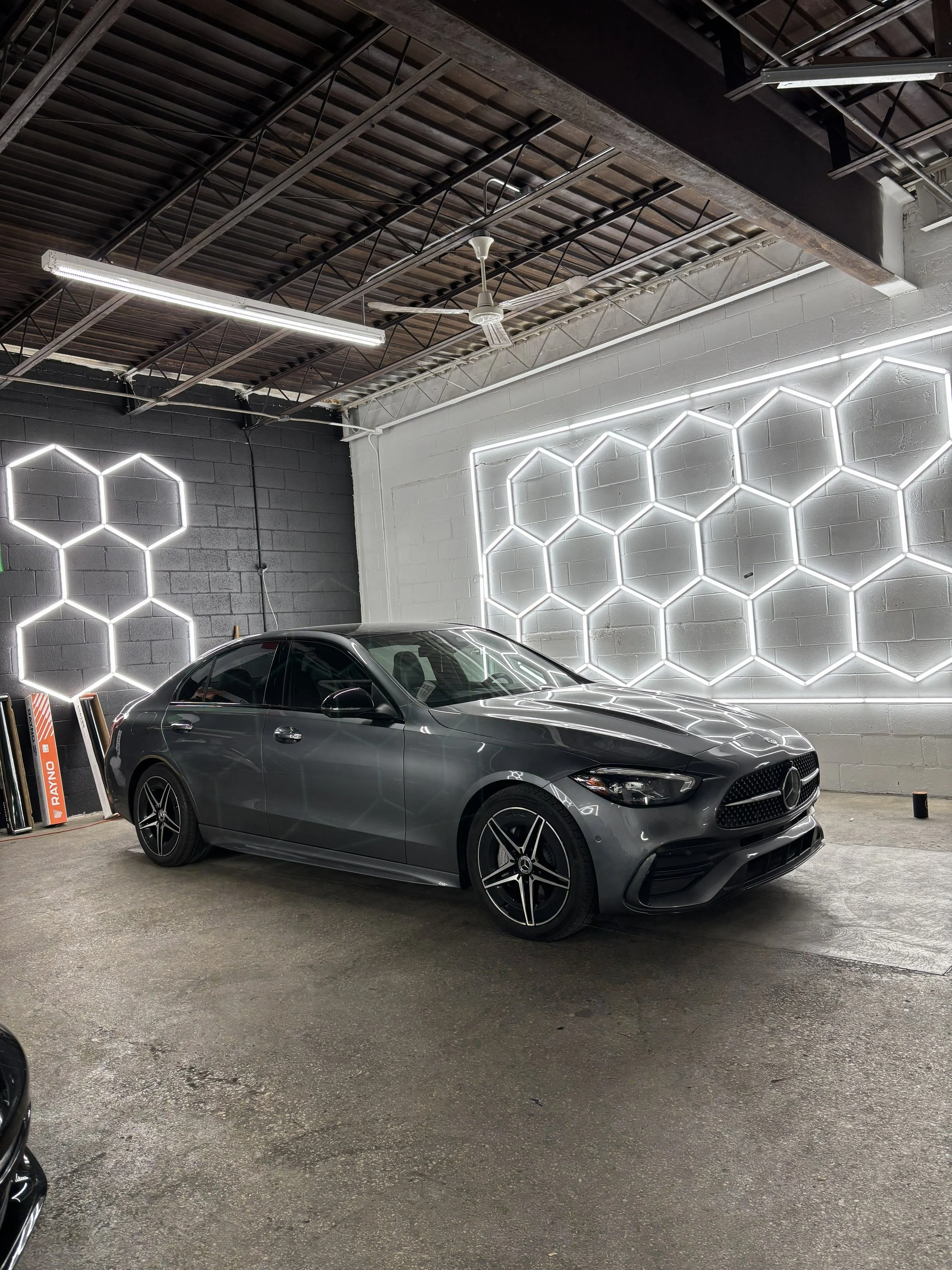 A dark gray Mercedes-Benz sedan parked inside a modern, industrial-style garage with illuminated hexagon-shaped wall decorations.