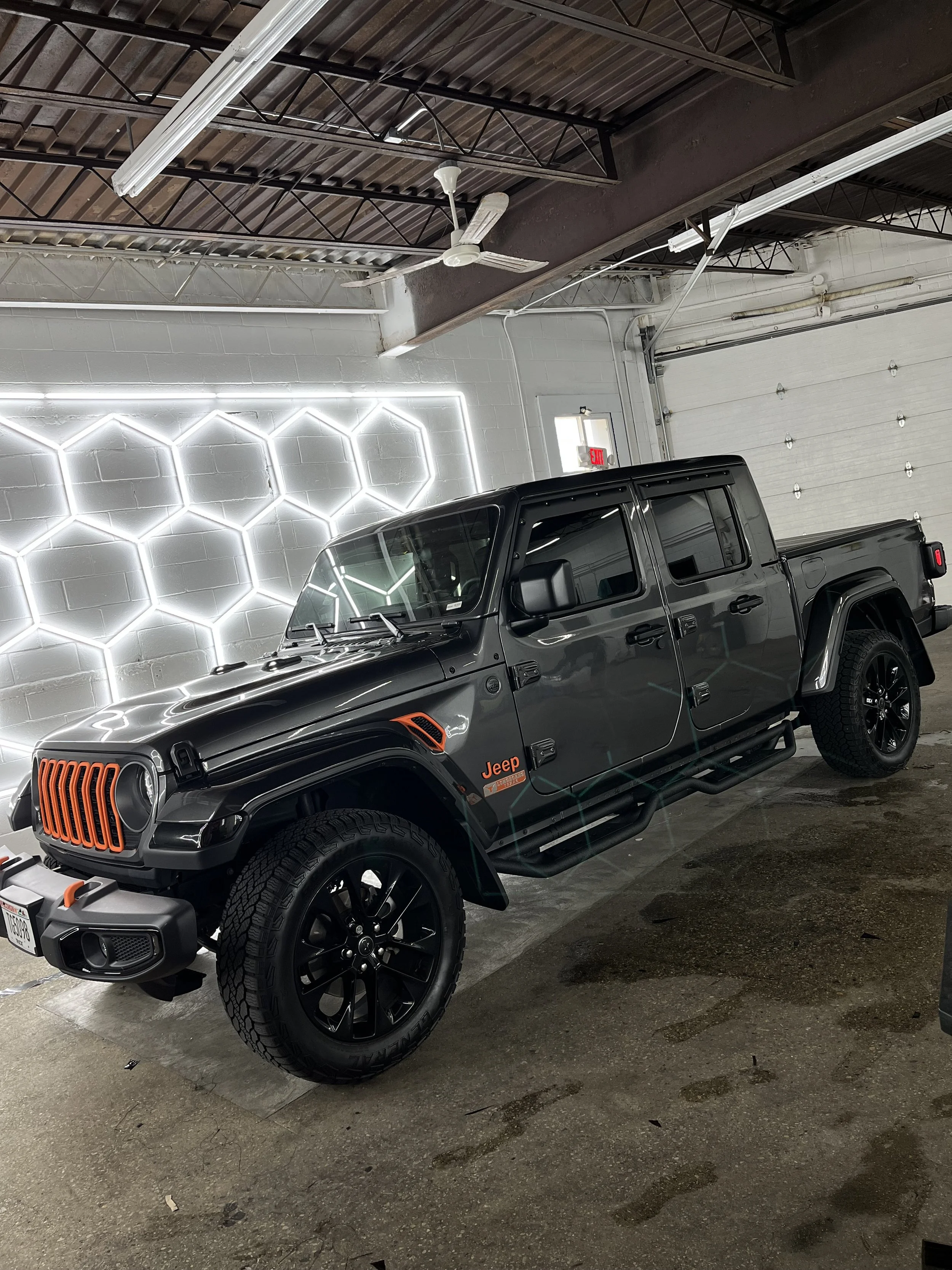 A black Jeep Gladiator truck parked inside a garage with white walls and hexagonal-shaped LED lights on one wall.