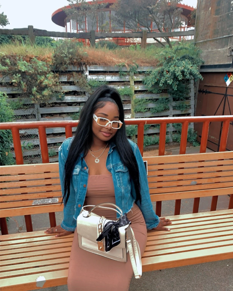 A woman with long black hair, wearing white glasses, a denim jacket, and a beige dress, sitting on a wooden bench outdoors with greenery and a building in the background.