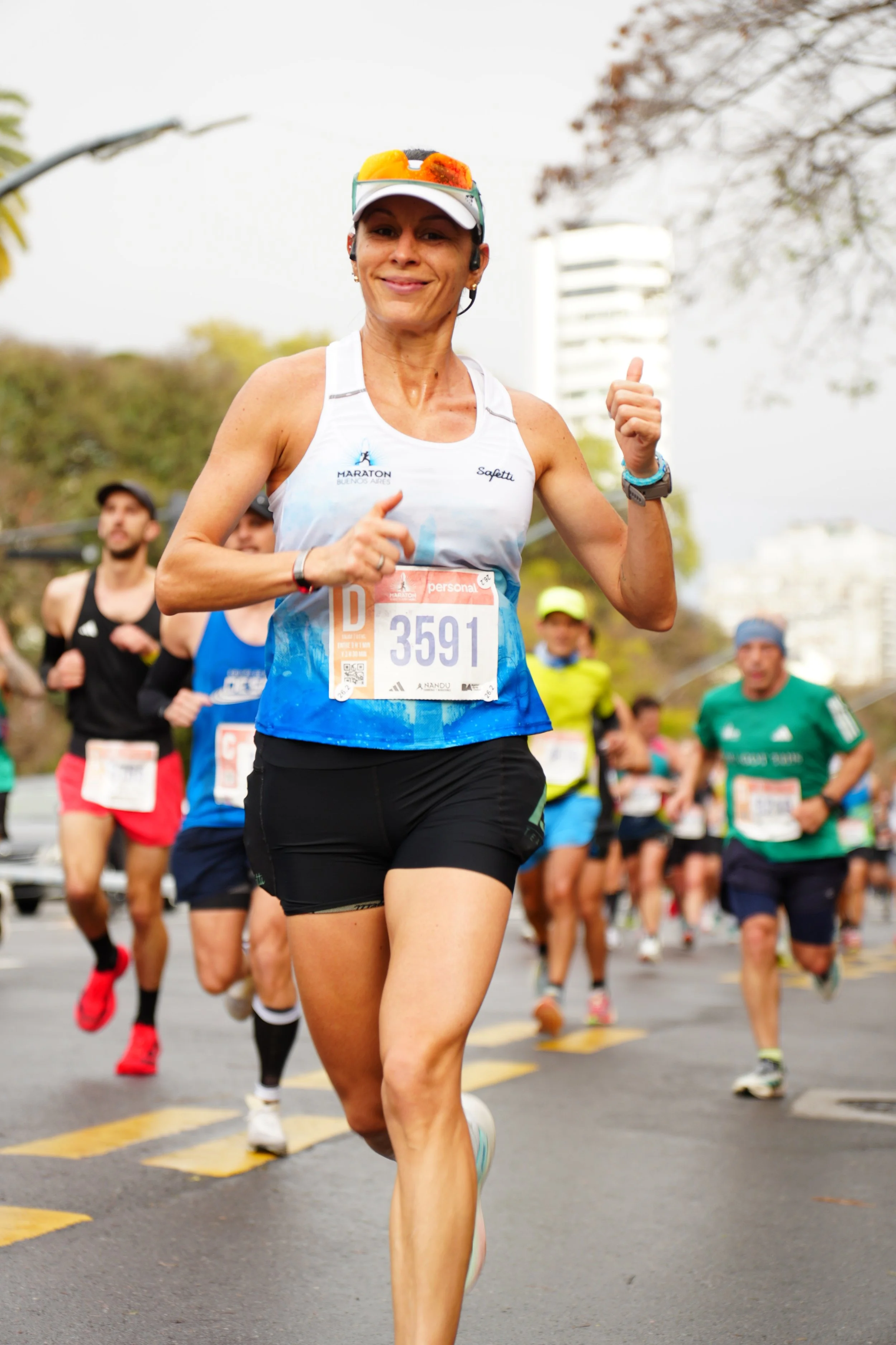 A woman running in a marathon race, wearing a white and blue tank top, black shorts, a white visor with sunglasses on it, and a race bib number 3591. Other runners are visible in the background.