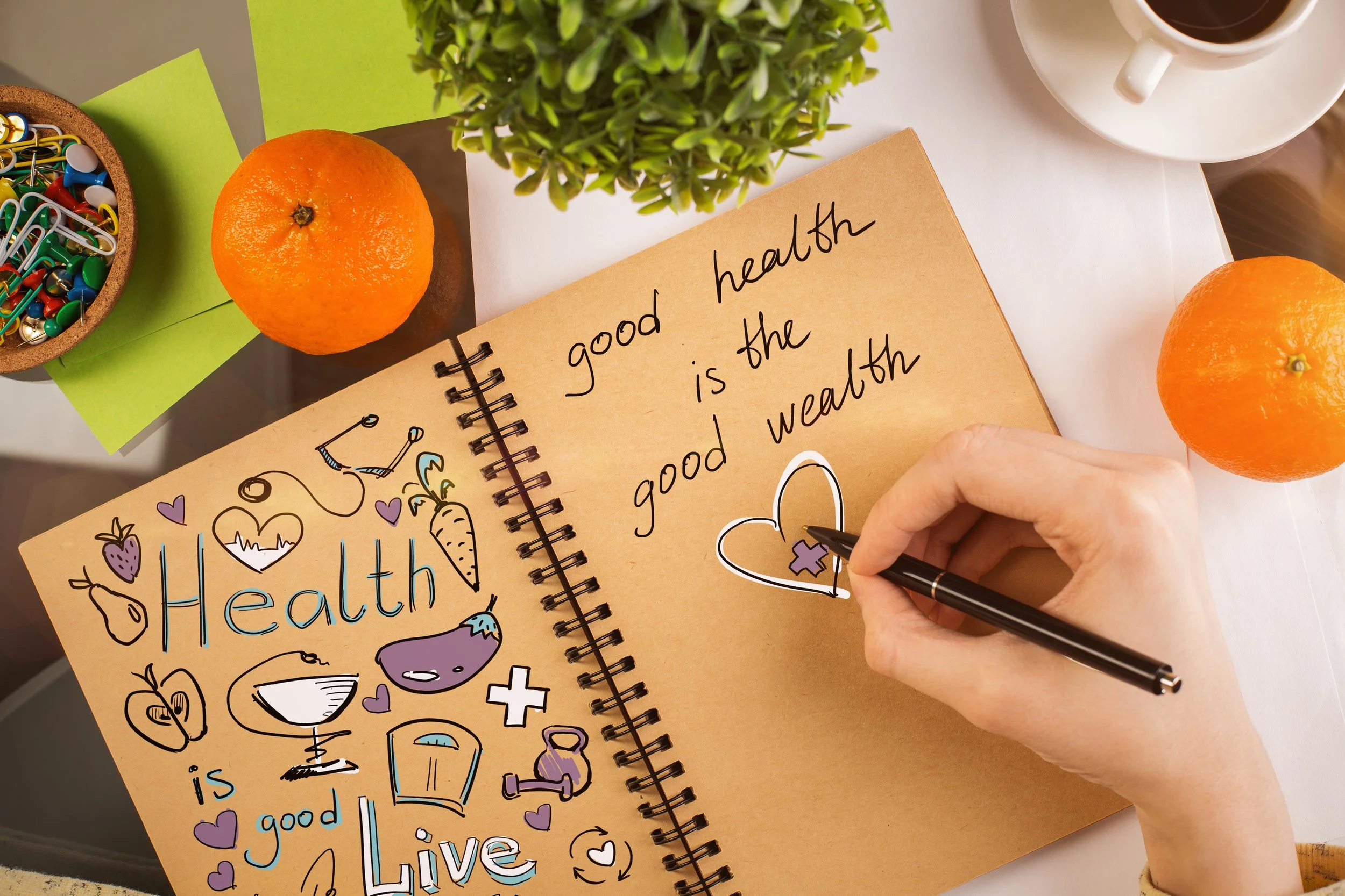 A person writing a message about health and good heart health in a notebook. The page contains handwritten words and a heart drawing with a puzzle piece. Surrounding the notebook are two oranges, a cup of coffee, a green plant, colorful paper clips, and a drawing of various healthy foods and symbols.