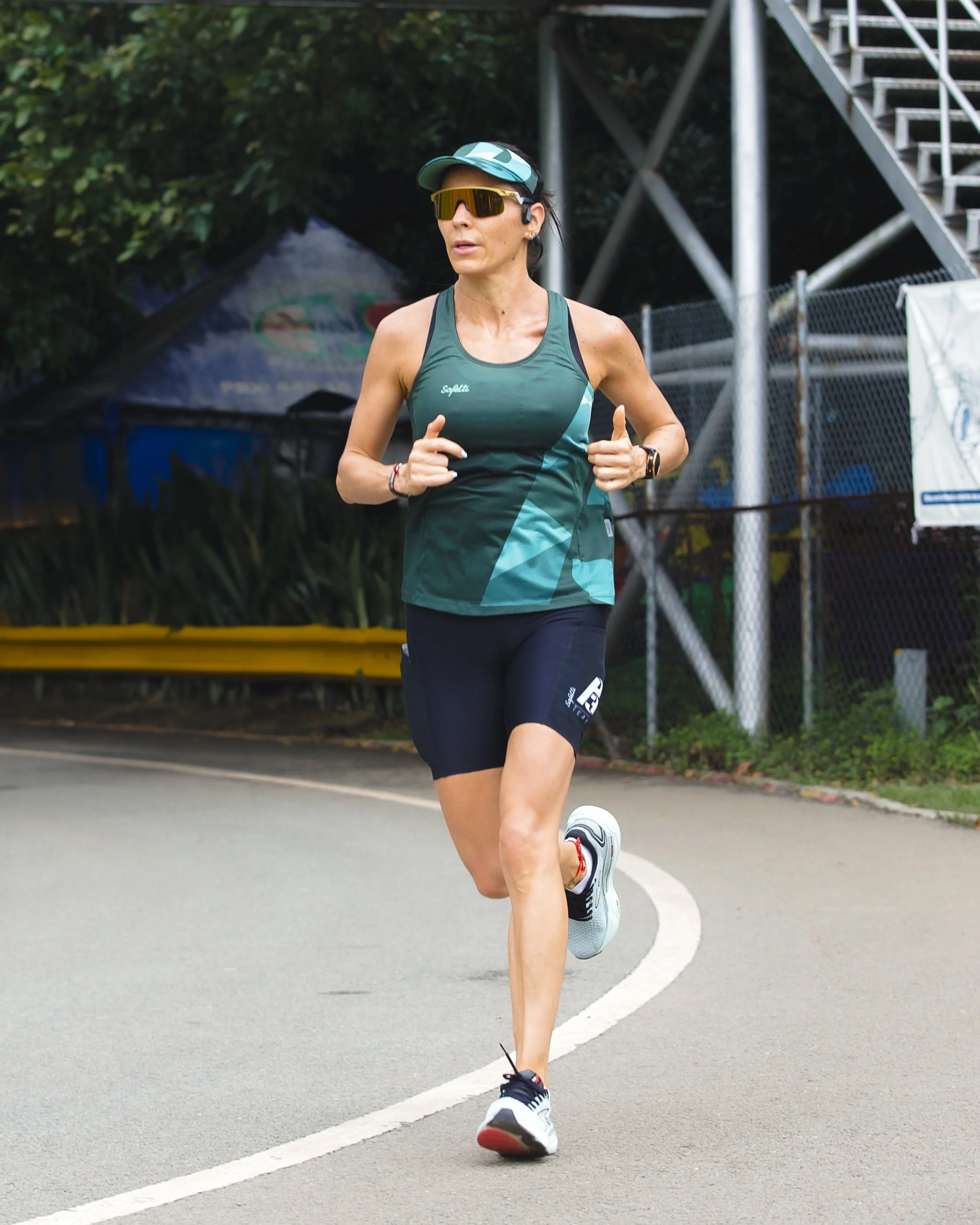 A woman running outdoors on a curved road, wearing athletic gear, sunglasses, and a cap.